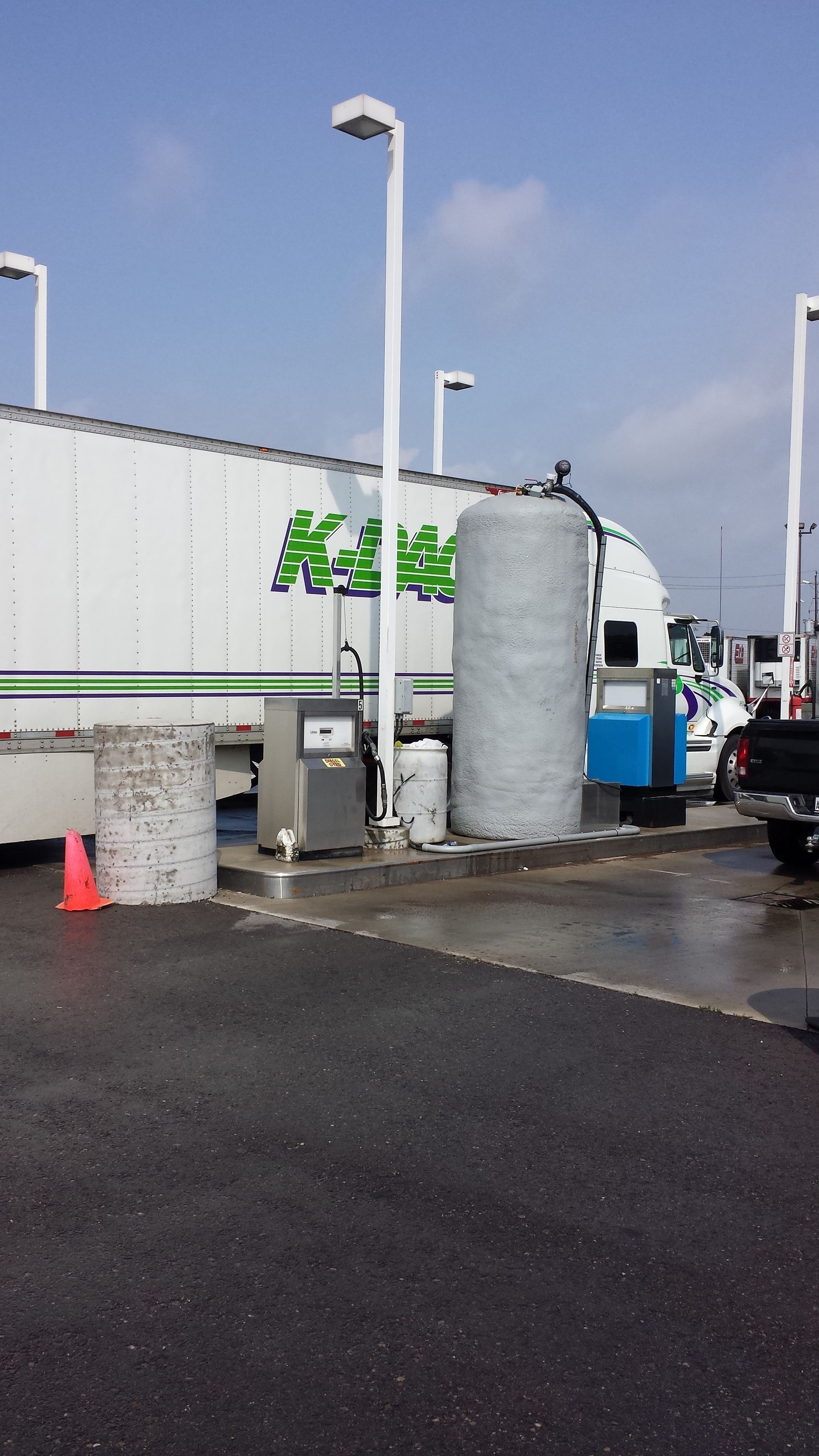 Semi-truck at a fuel pump with a large white tank. Green logo on the trailer, bright blue sky, asphalt ground.