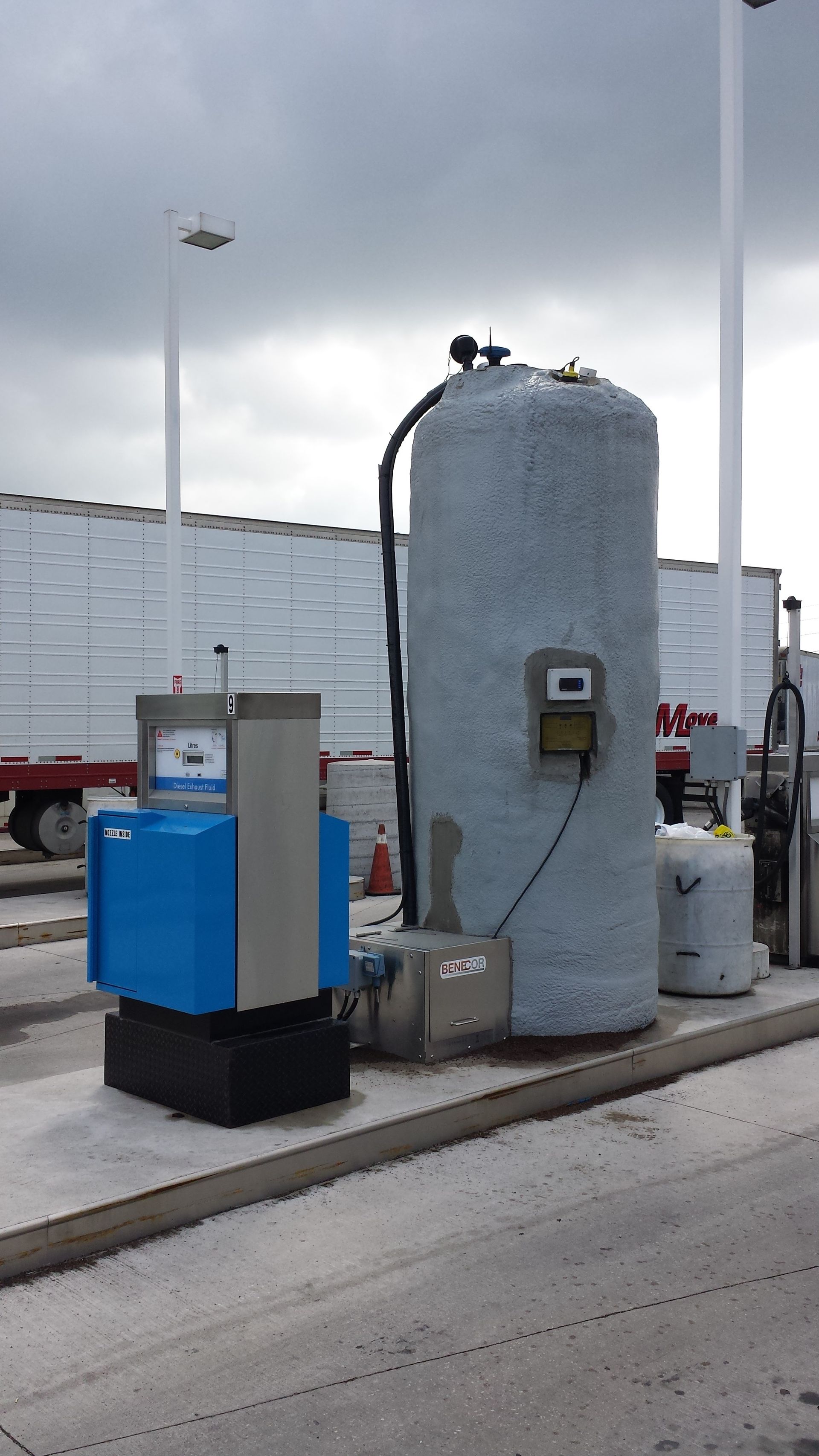 Fueling station with a blue pump, tall tank, and semi-truck trailers in the background under a cloudy sky.