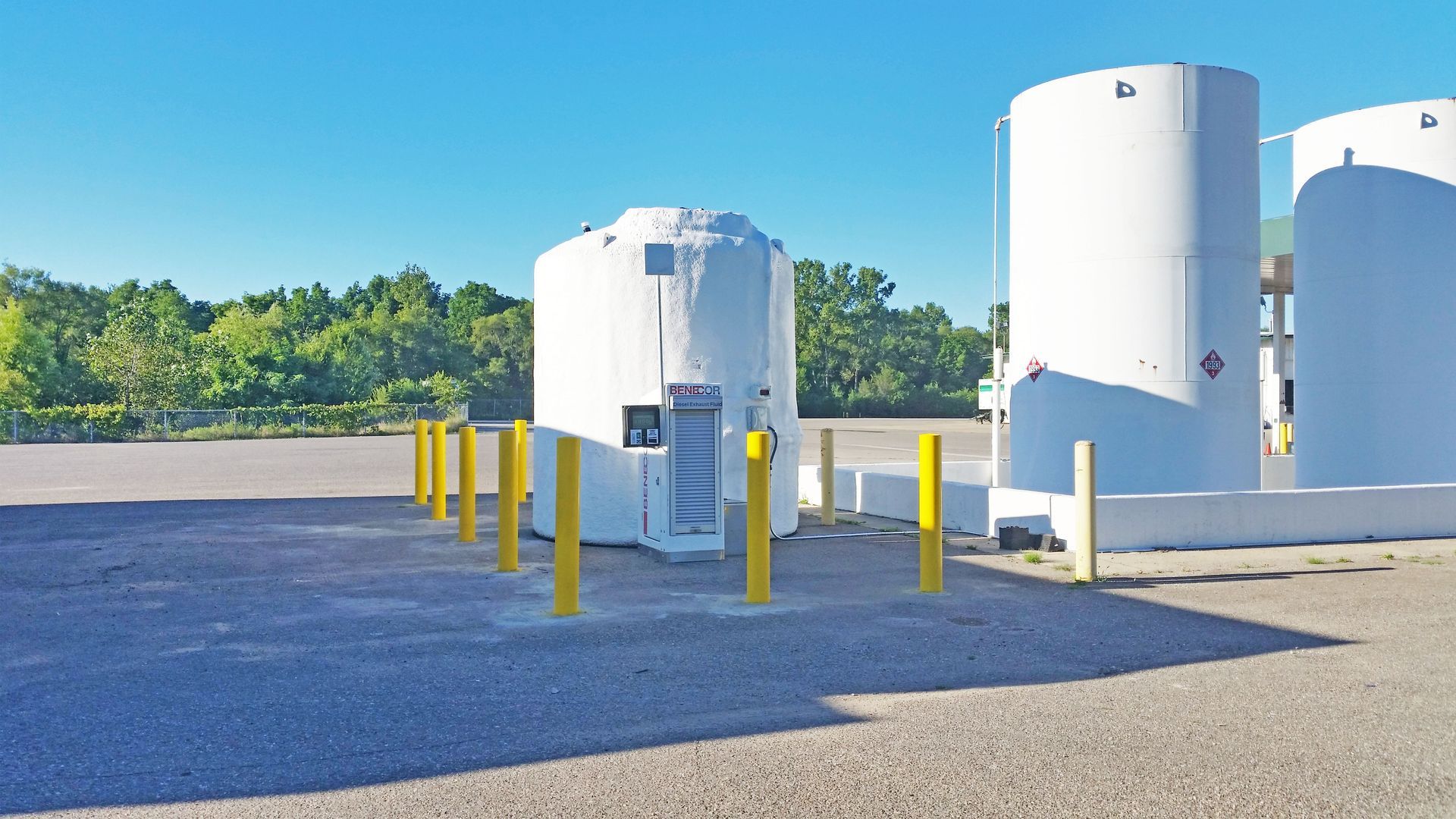 White industrial tanks on a paved lot with yellow bollards, trees in the background, and a bright blue sky.