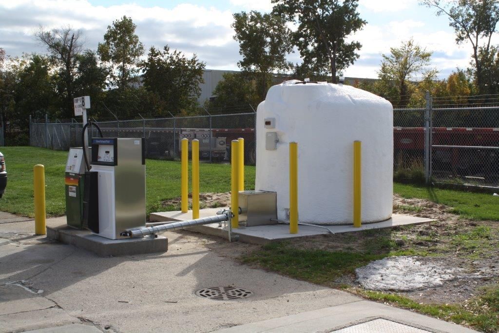 Gas pump and white tank. Yellow posts protect them. Outside, near fence and trees.