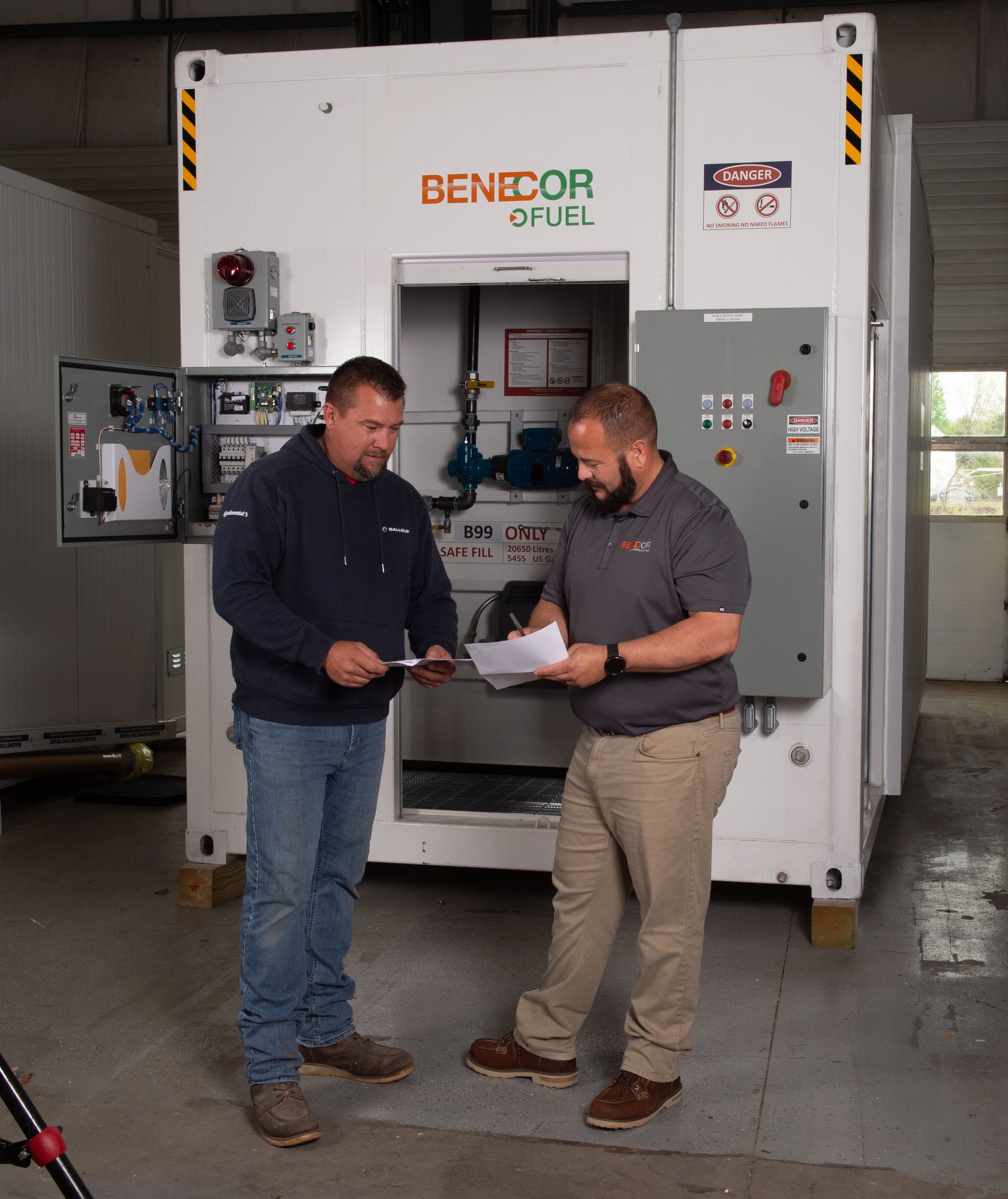 Two men review paperwork near a large white industrial machine, indoors. One man points.
