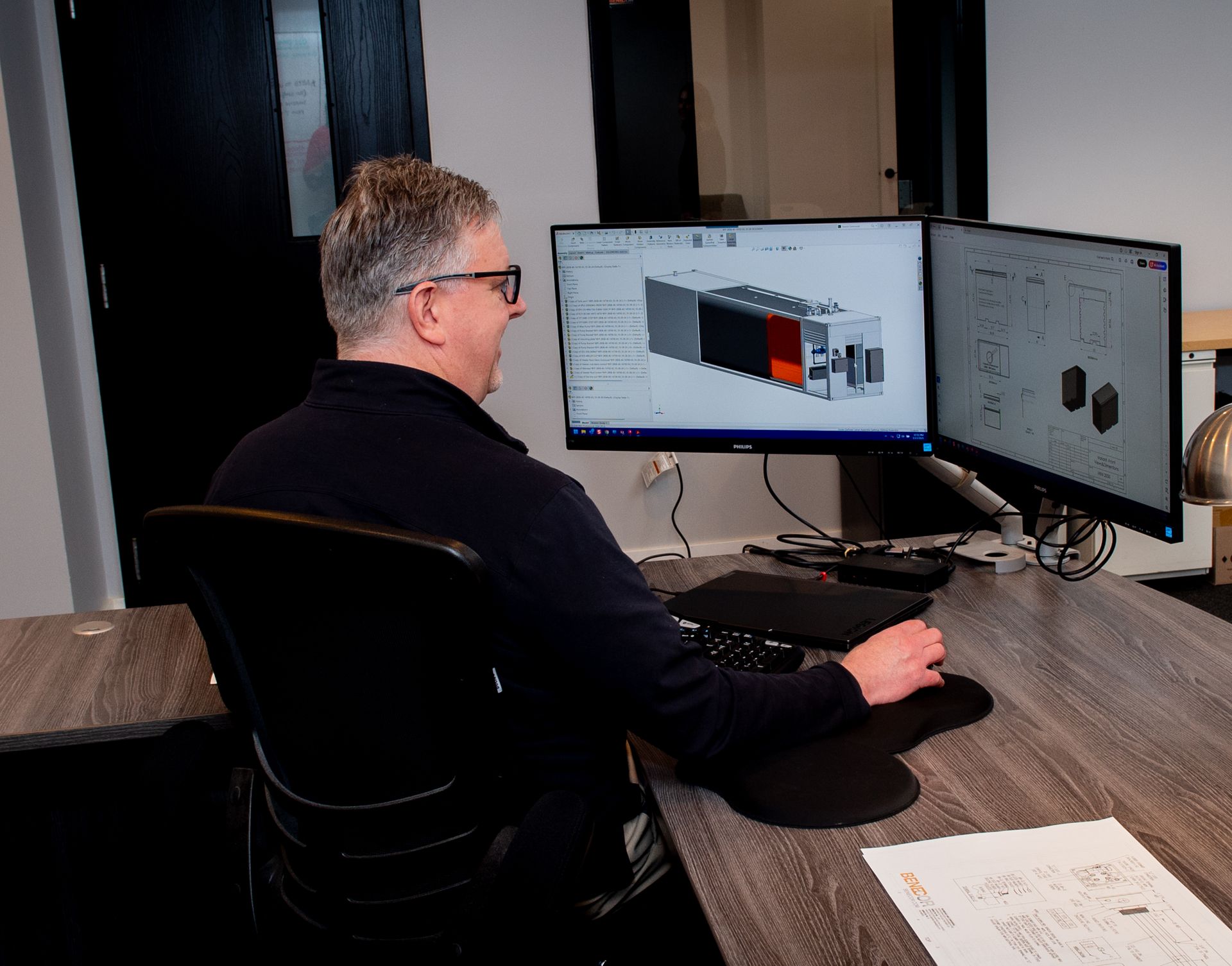 Man using computer with dual monitors; drafting design at desk.