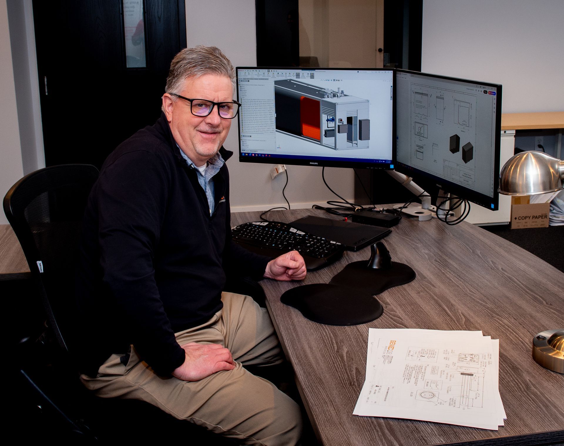 Man smiling at a desk with dual monitors showing 3D design and diagrams. He's wearing glasses in a dark office.