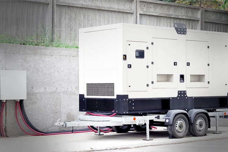 White generator on a trailer, next to a wall, with power cables connected.