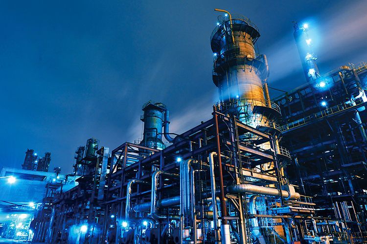 Oil refinery at night, illuminated by blue lights. Pipes and towers dominate the scene against a dark sky.