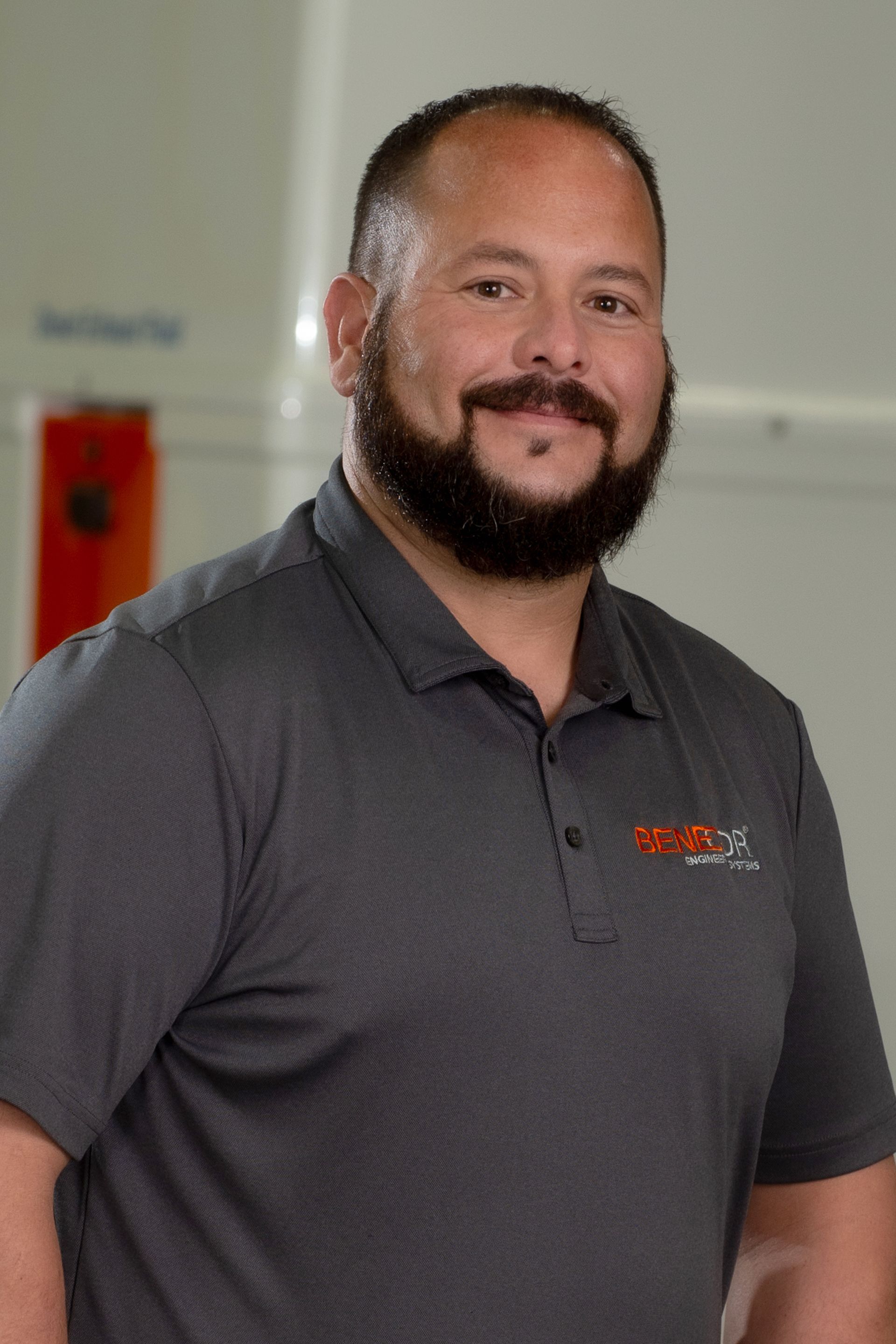 Man with a beard smiling, wearing a gray polo shirt with a logo; blurred background.