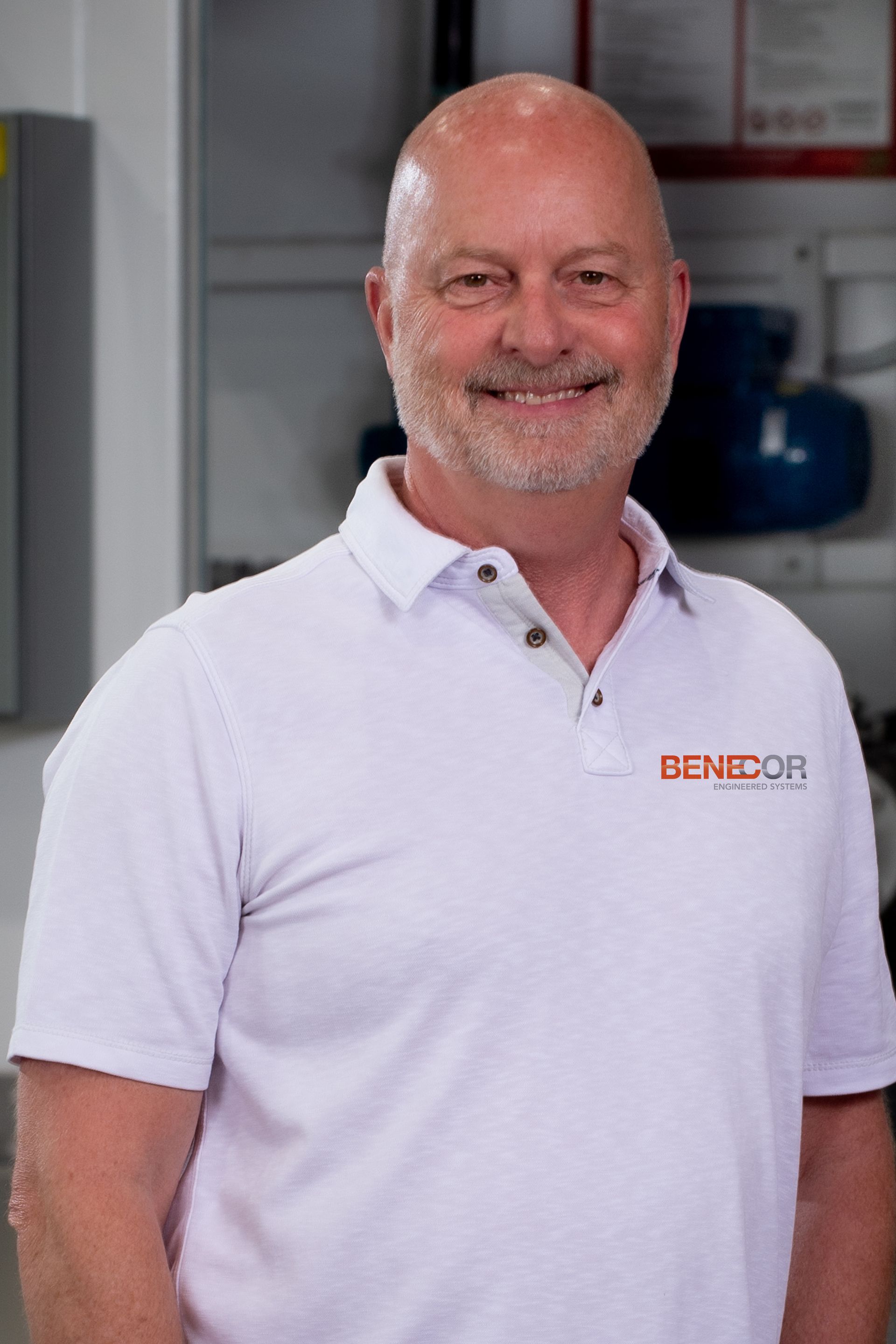 Bald man in white polo shirt smiles, in a factory setting.