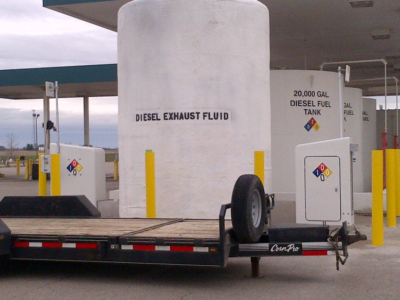 A white diesel exhaust fluid tank at a fuel station, with a flatbed trailer in the foreground.