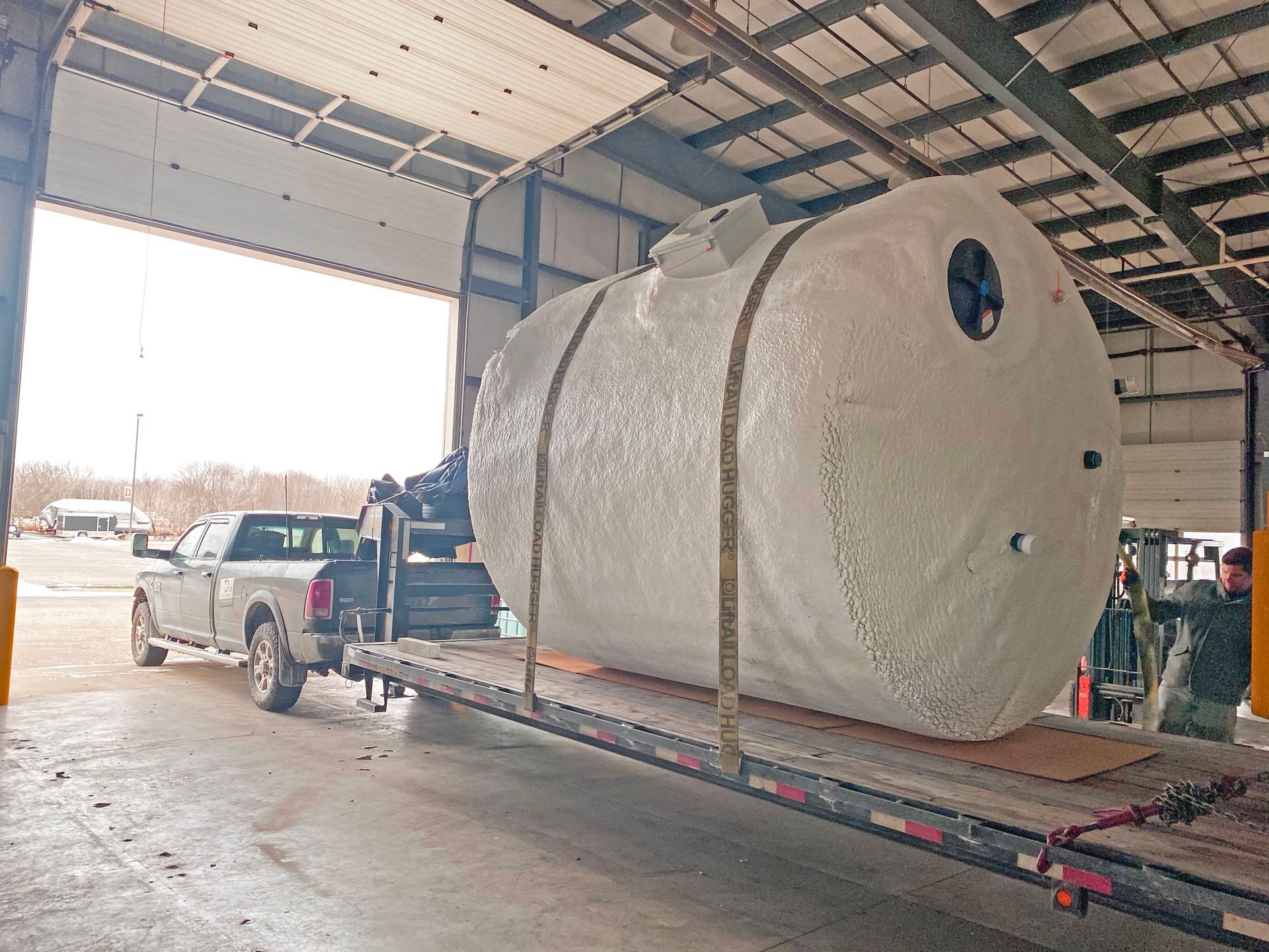A large white tank is being loaded onto a flatbed trailer from a loading dock by a truck and crane.
