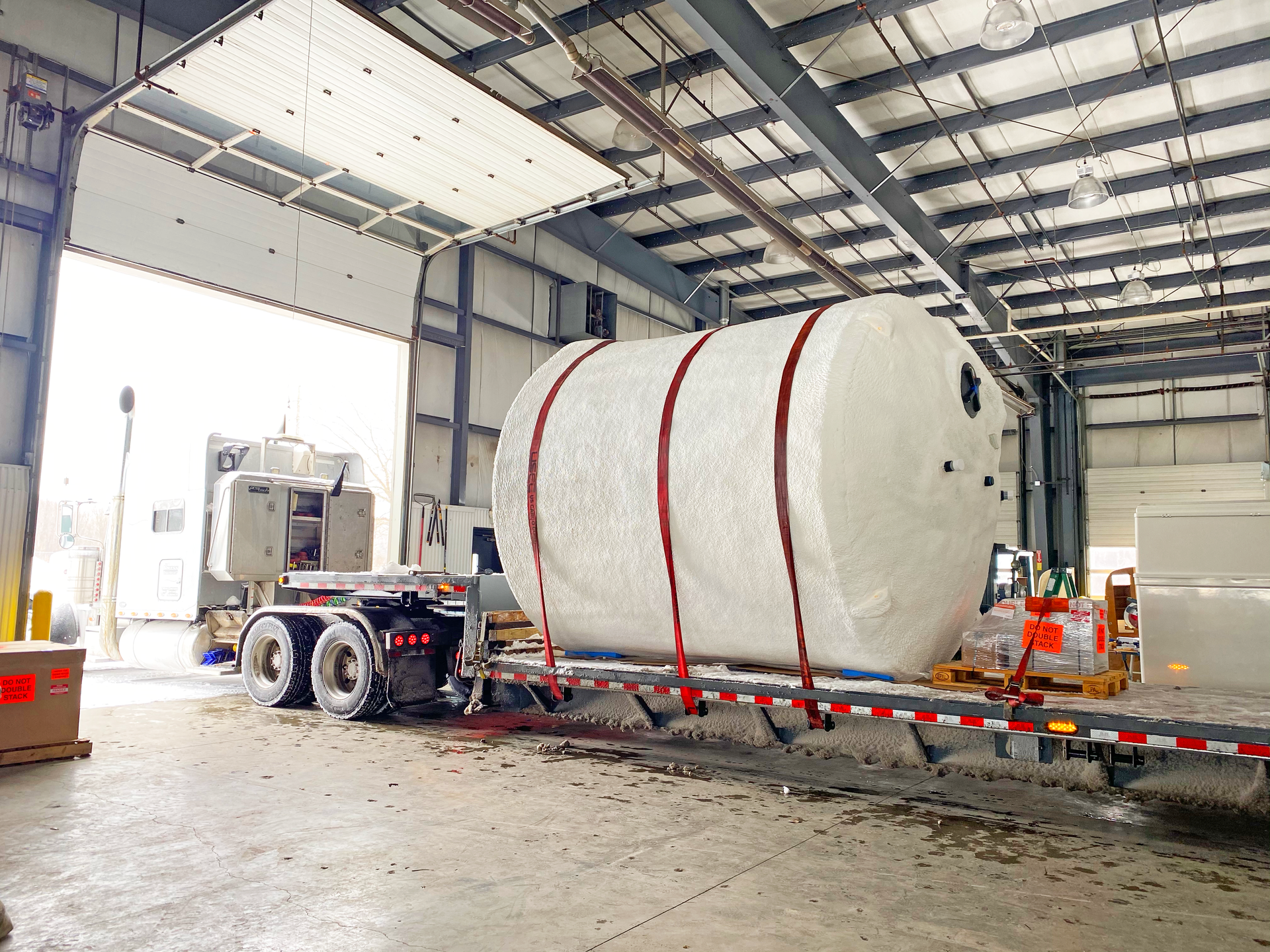 A semi-truck trailer carrying a large, wrapped cylindrical tank is inside a warehouse.