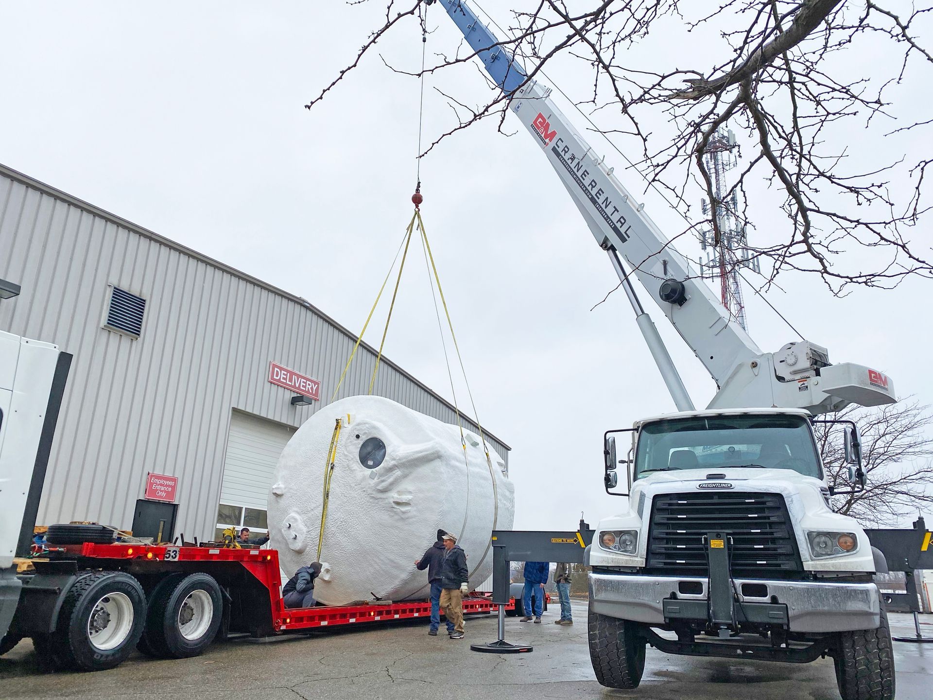 Crane lifting a large, white, wrapped tank from a flatbed truck next to a building. People are nearby.