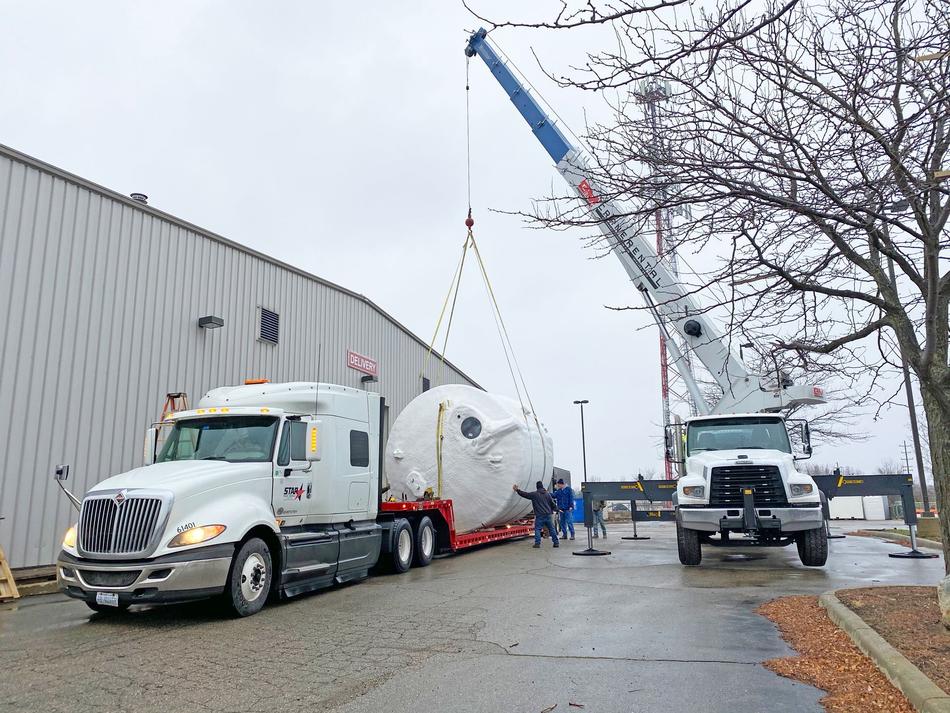 Truck transporting a large white tank, being lifted by a crane near a building on a cloudy day.