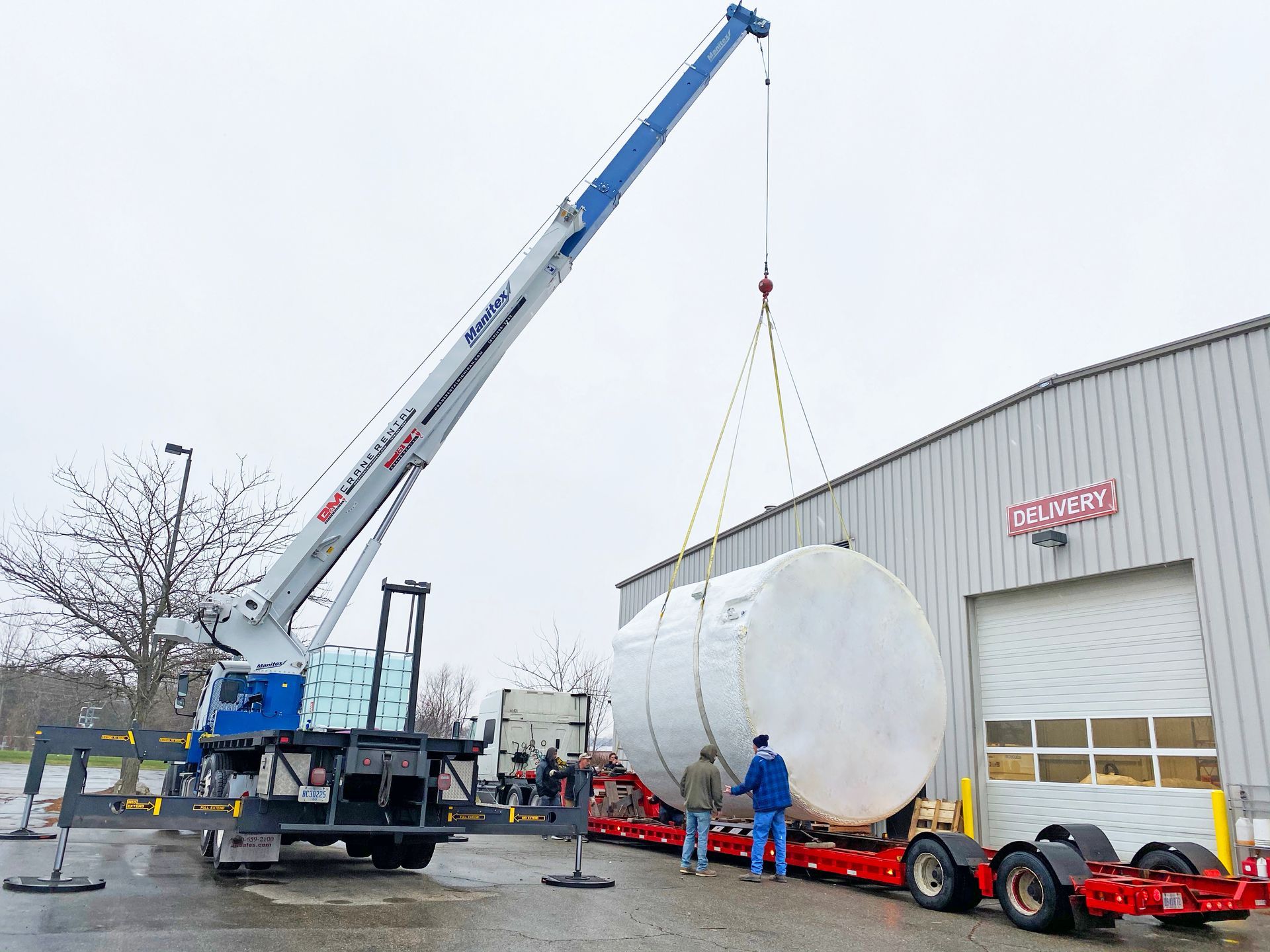 A crane lifting a large, white tank from a red trailer near a building; two men watch.