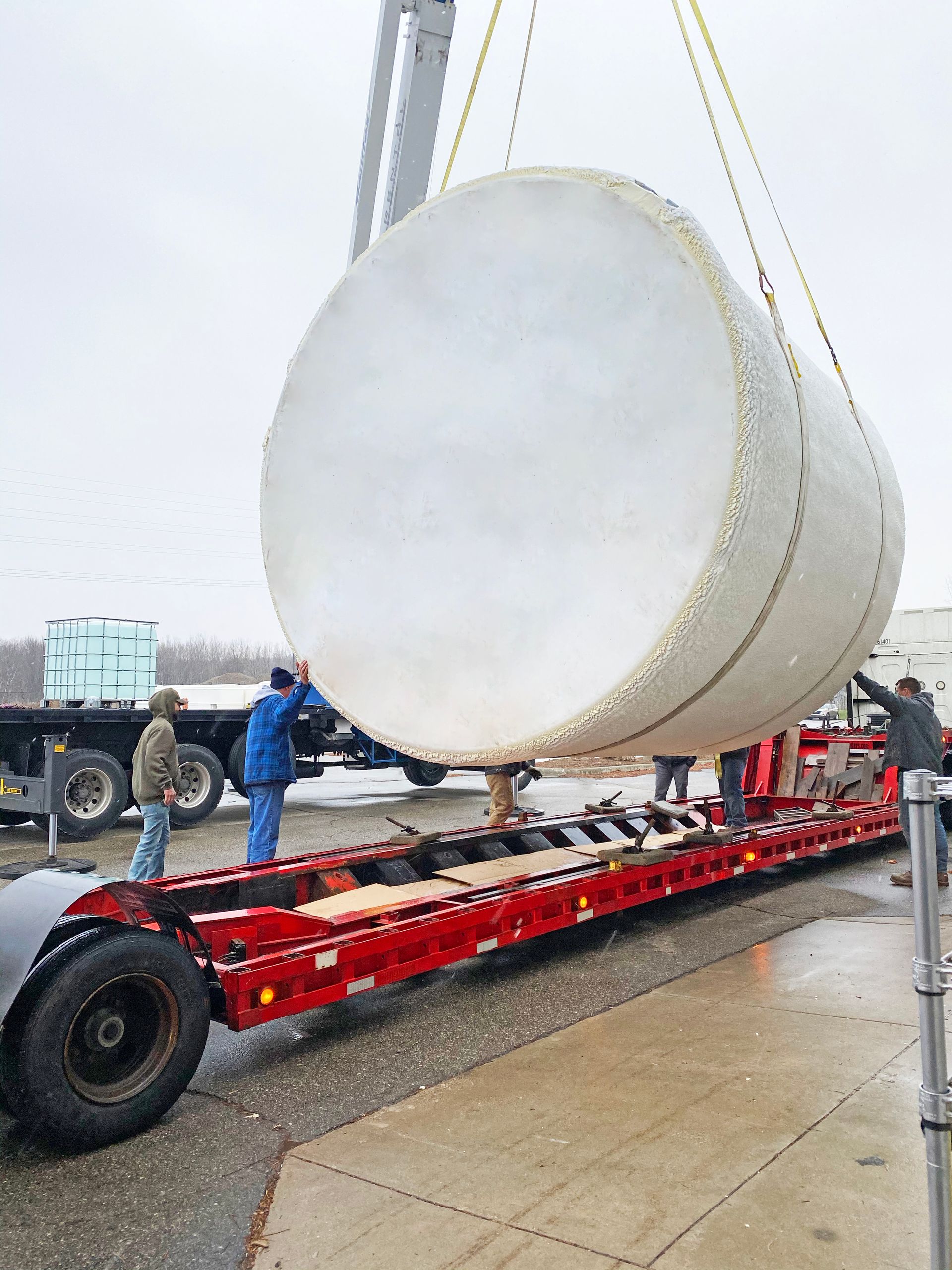 Large cylindrical tank being lifted onto a flatbed trailer by a crane. Workers are guiding it.