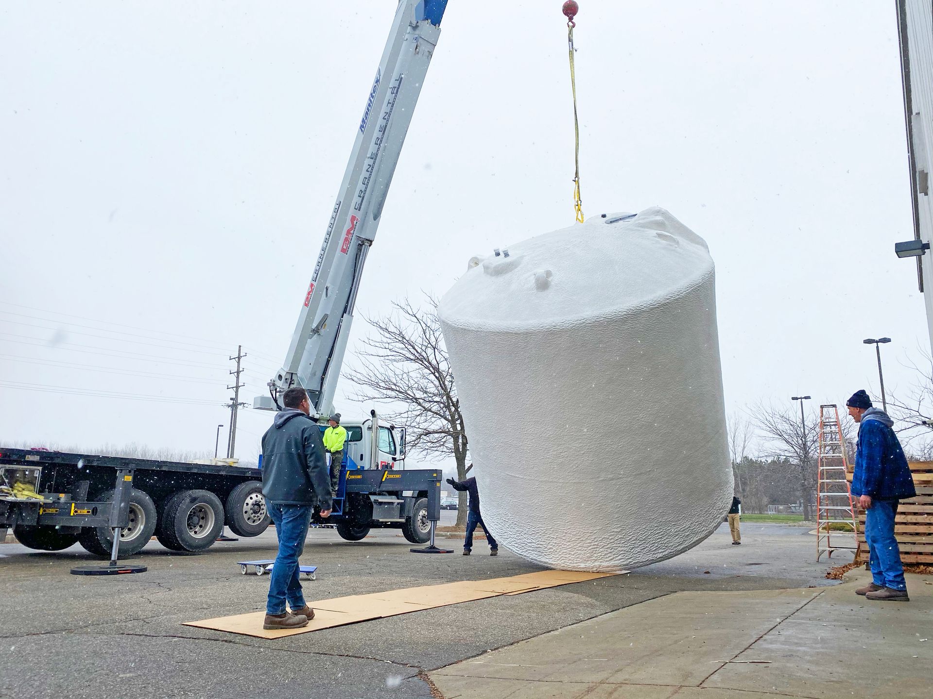 A crane lifts a large, cylindrical, white tank. Workers watch in a gray setting.