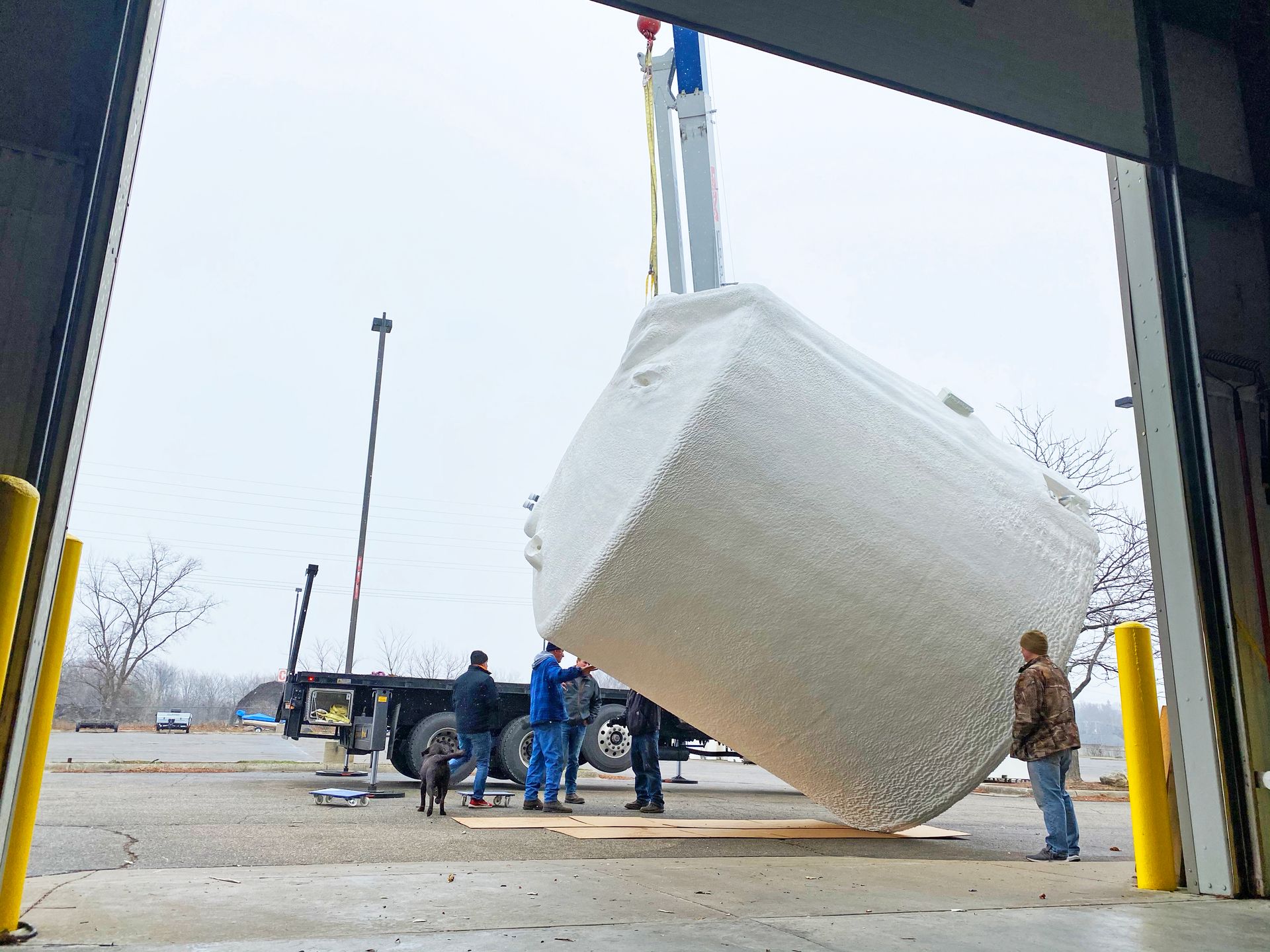 Crane lifts a large, white object wrapped in plastic, near a truck and warehouse.