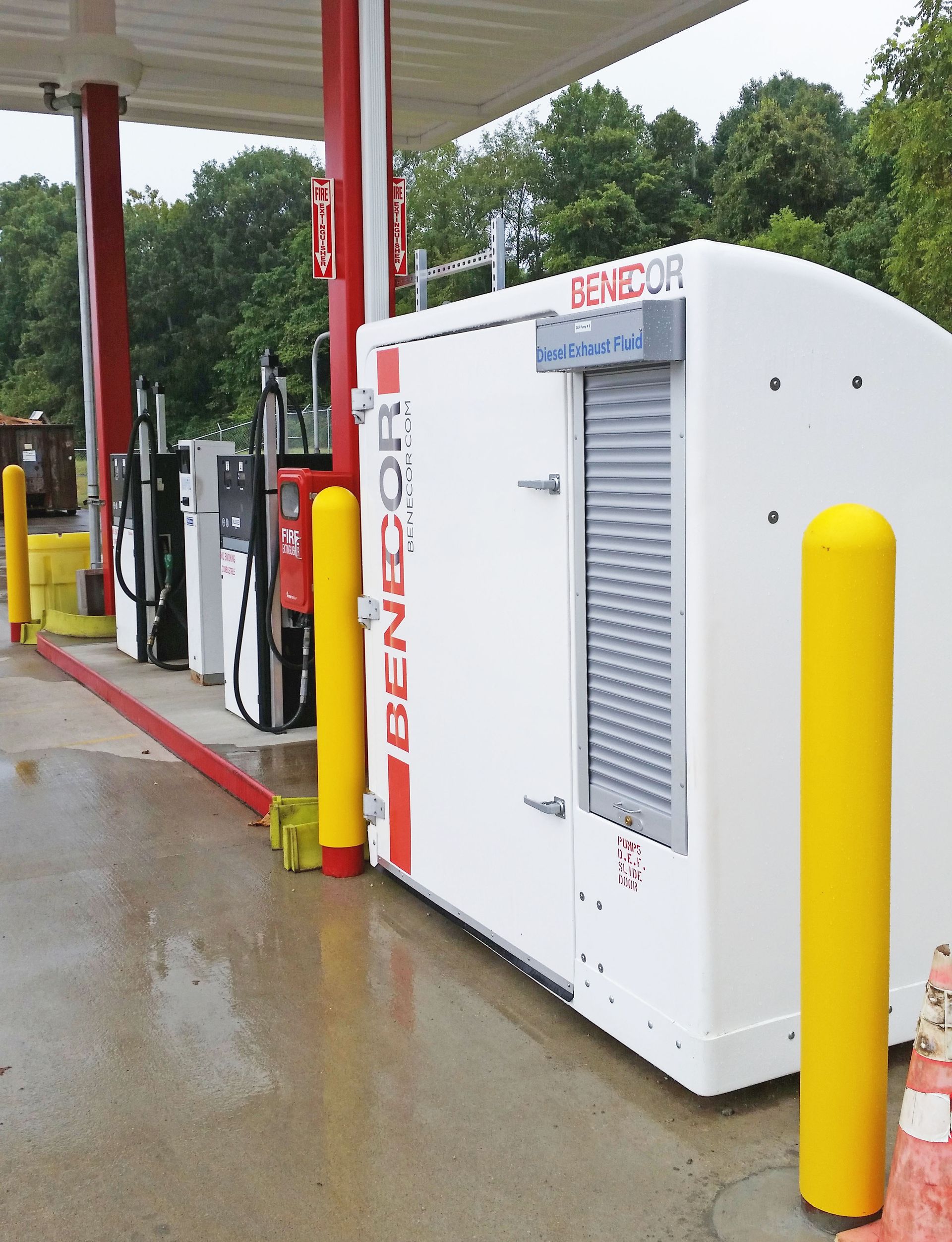 A white BENECOR machine at a gas station next to yellow poles and gas pumps.