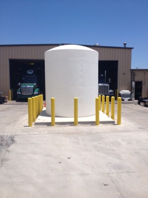 White cylindrical tank on a concrete pad, surrounded by yellow bollards, in front of a building with a truck visible inside.