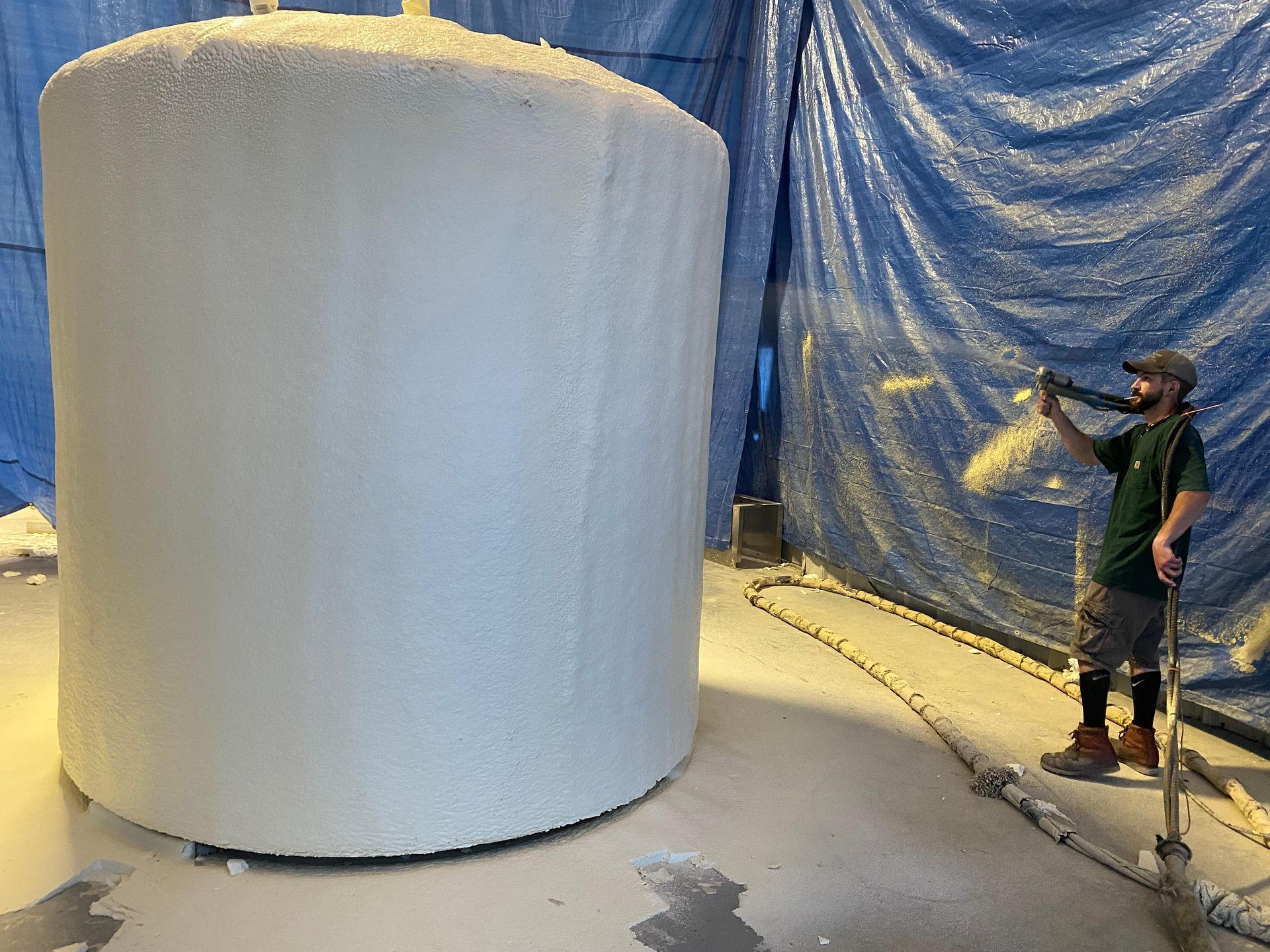 Person spraying a large, cylindrical structure covered in white, within a blue-draped room.