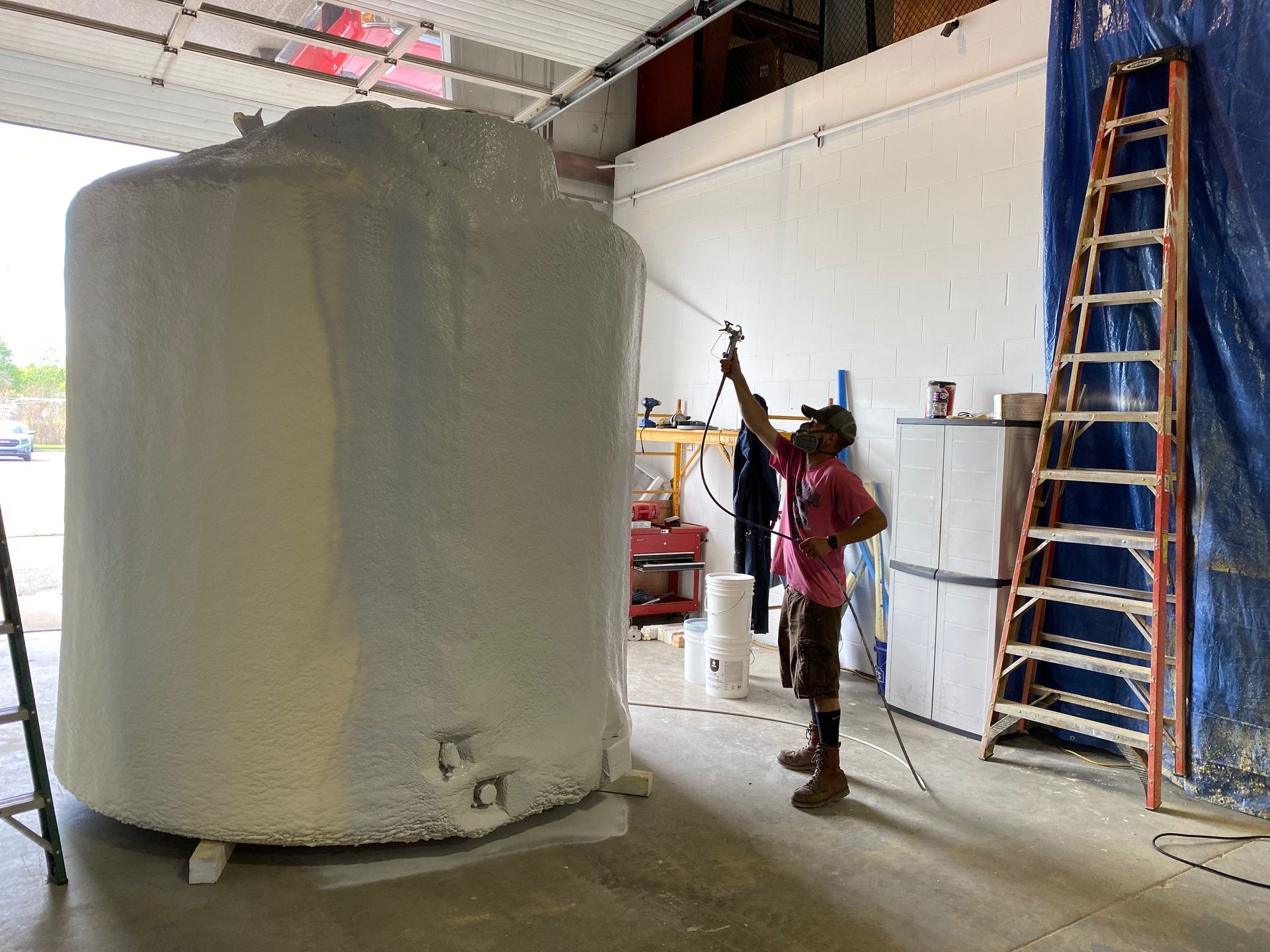 Man sprays paint on a large white tank in a garage. Ladder and work supplies visible.