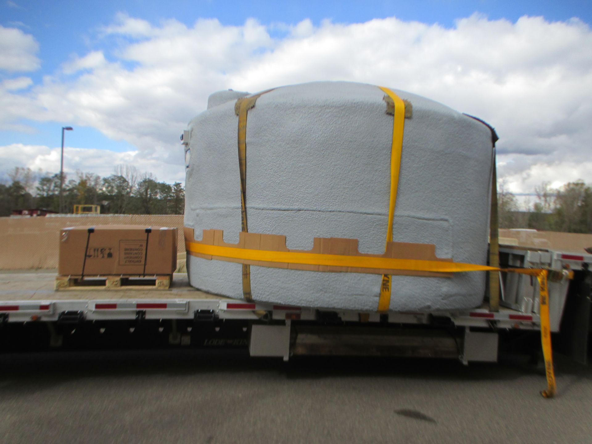 A large gray water tank strapped to a flatbed truck, with a smaller box on a pallet nearby.