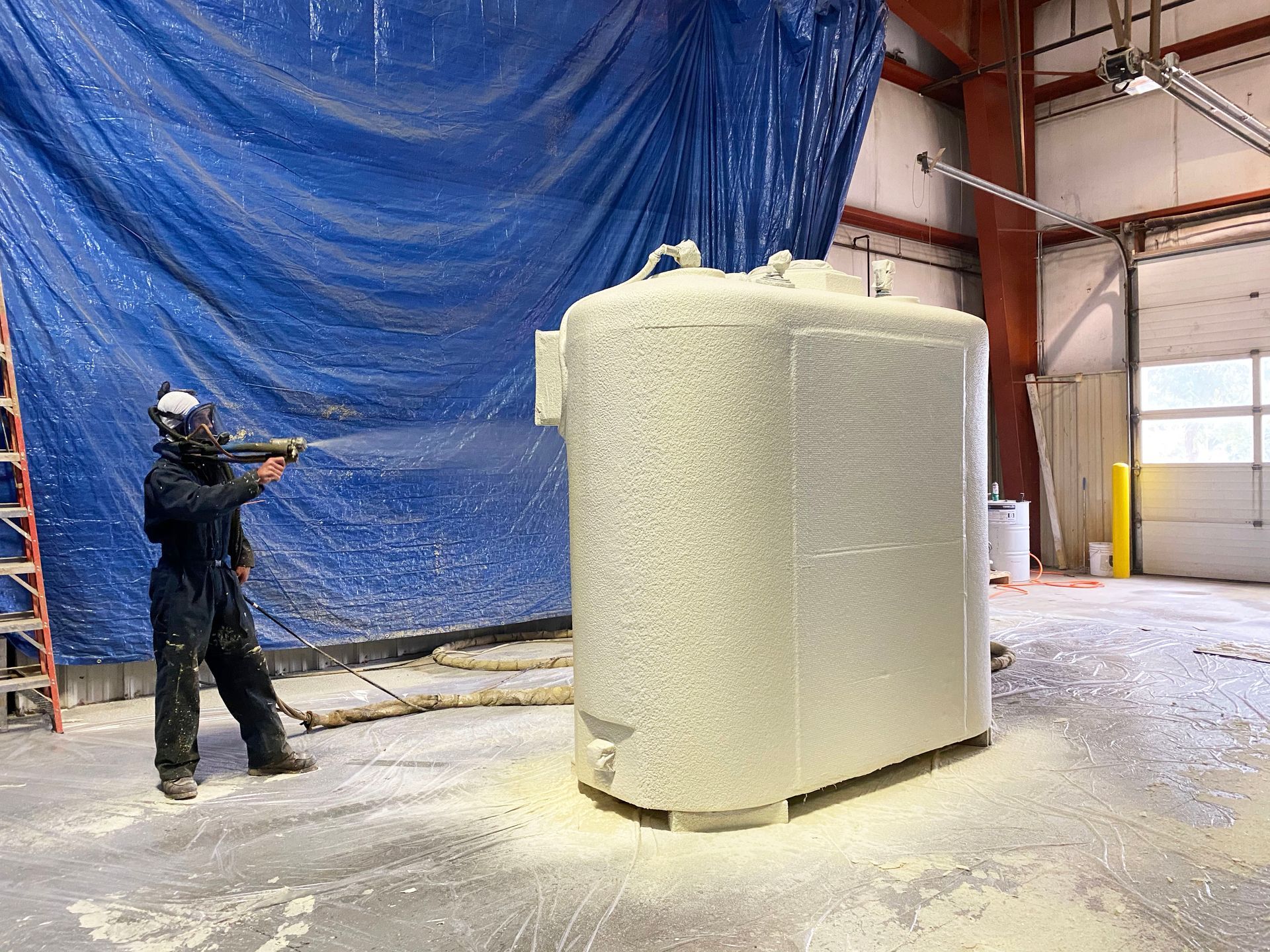 Person in protective gear spraying foam insulation on a large, light-colored tank inside a warehouse.
