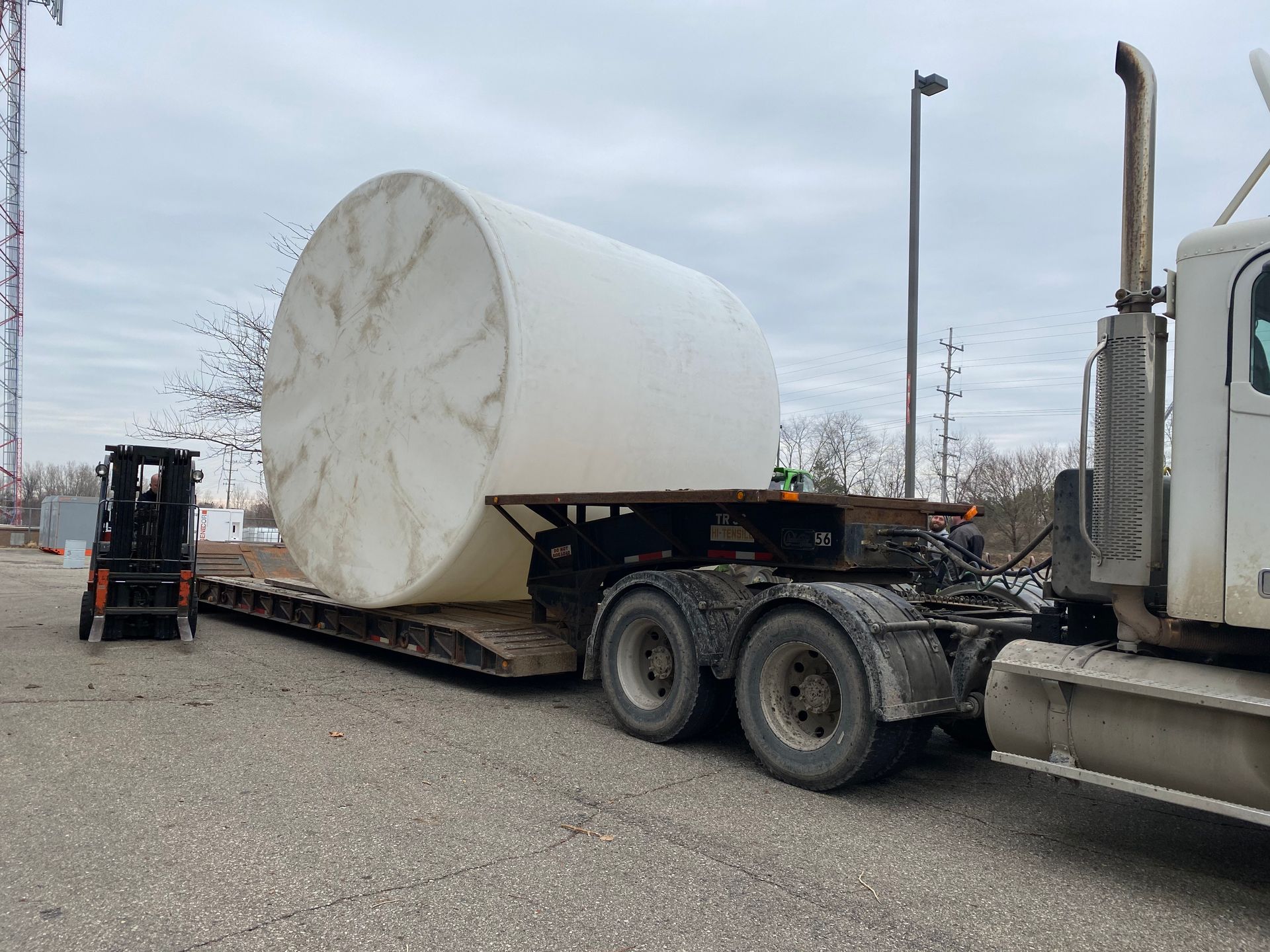 Large white cylindrical tank on a flatbed truck, with a forklift nearby and a truck cab to the right.