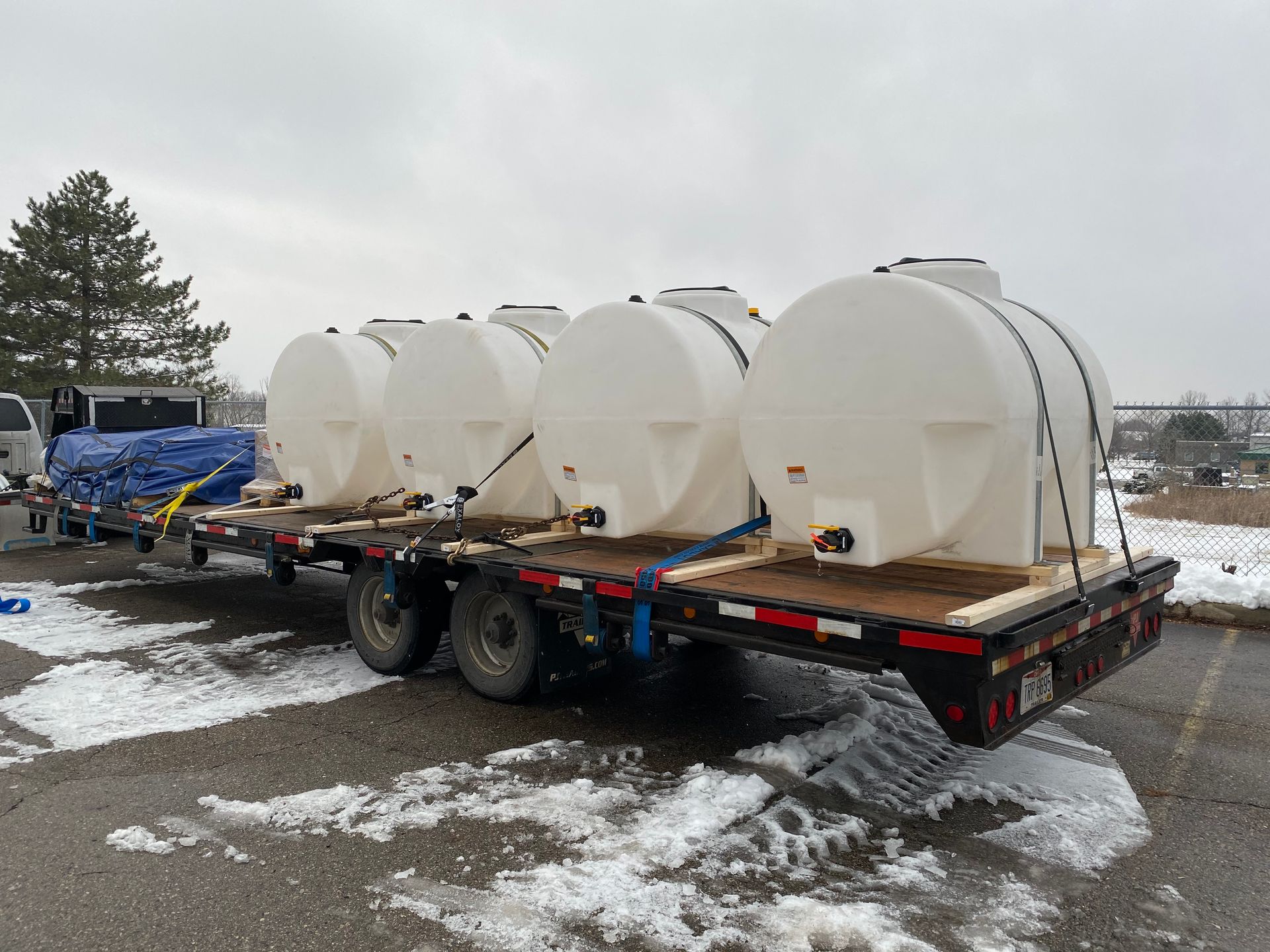 Four large white tanks secured on a flatbed trailer; snowy asphalt.