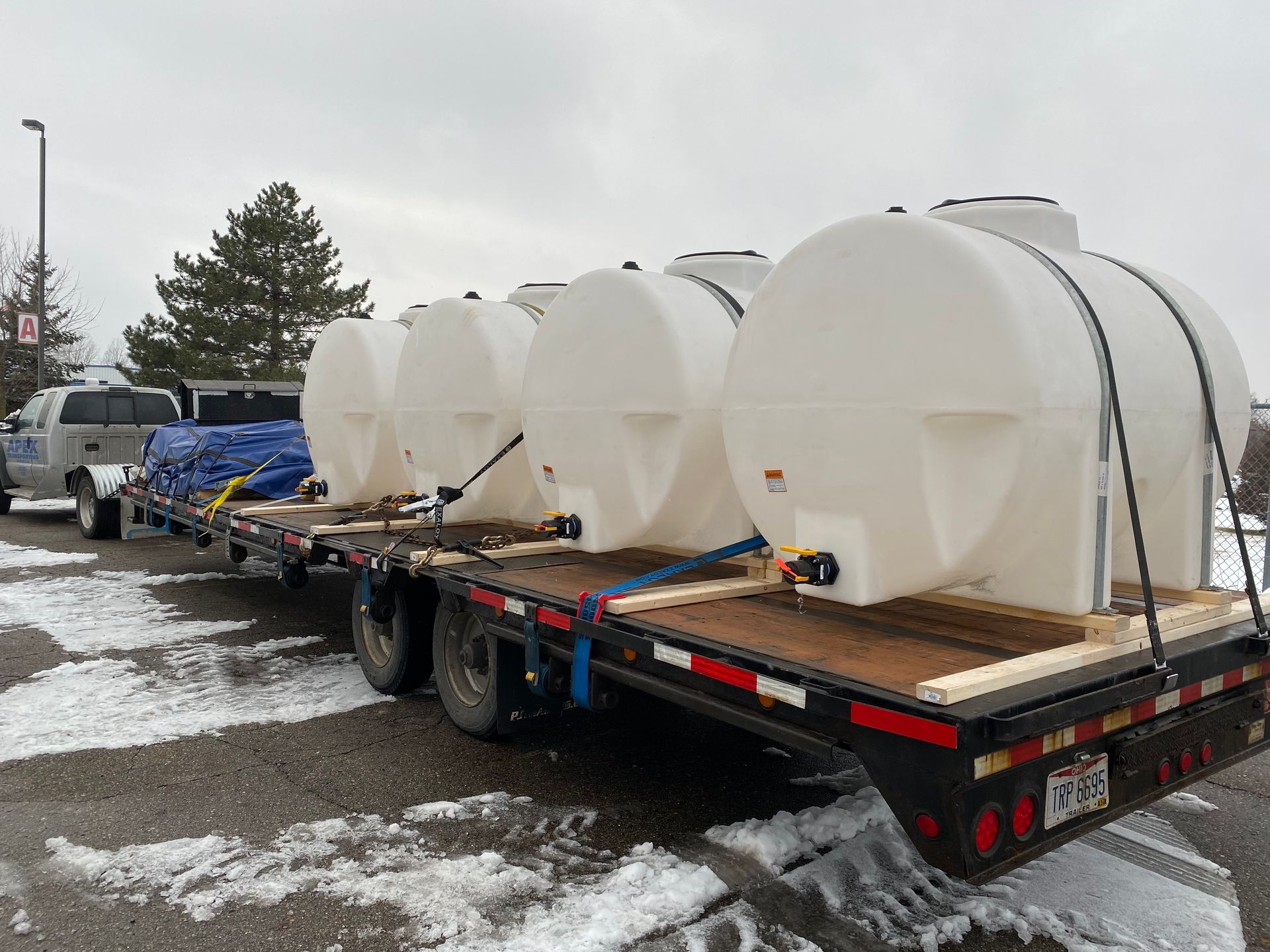 White water tanks secured on a flatbed trailer, possibly for transport. Winter setting, overcast sky.
