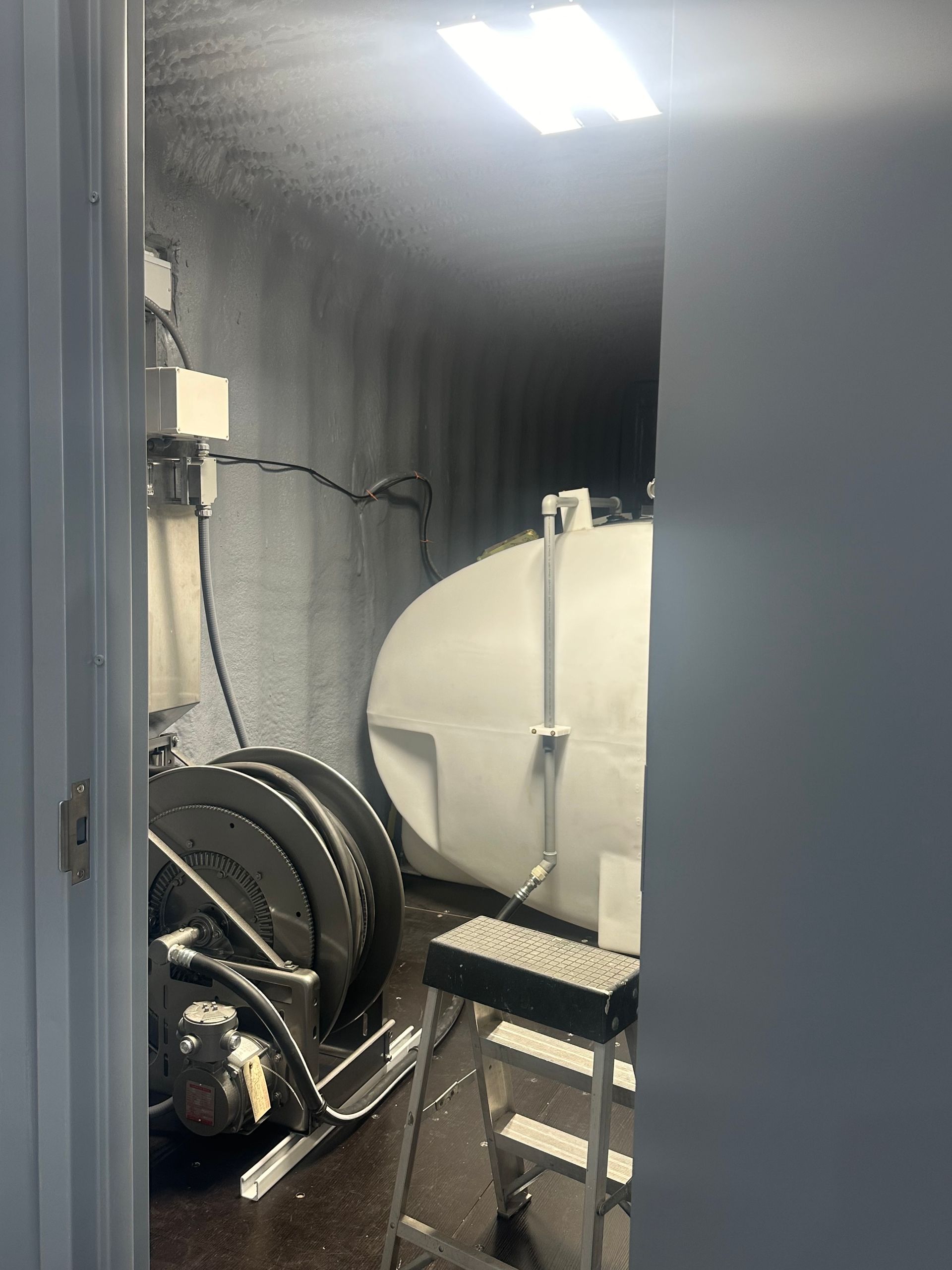 A small, dark room with industrial equipment: a large tank, hose reel, and a stool.