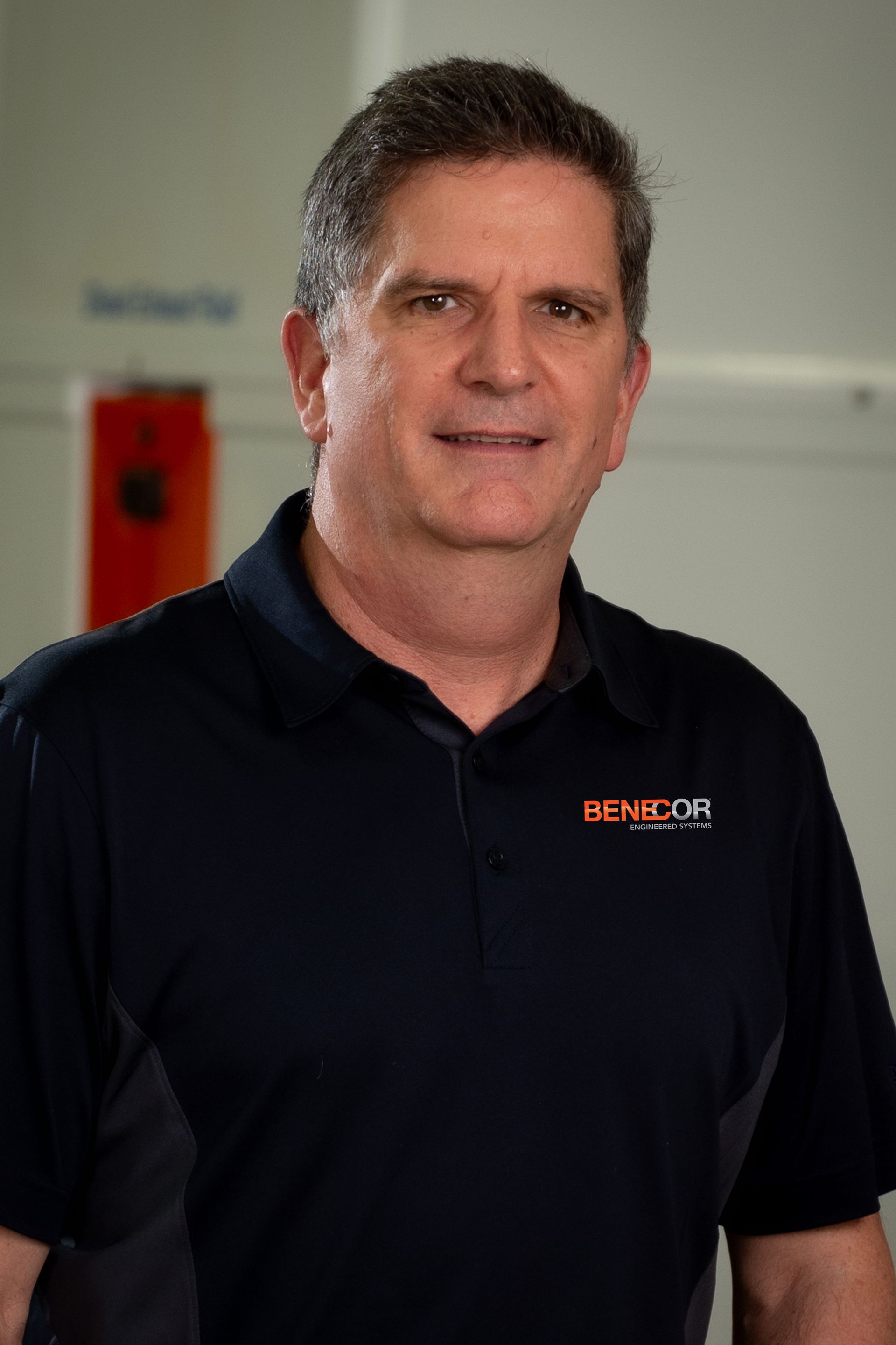 Man in a dark blue polo shirt smiles; the shirt has a logo. Indoor setting.