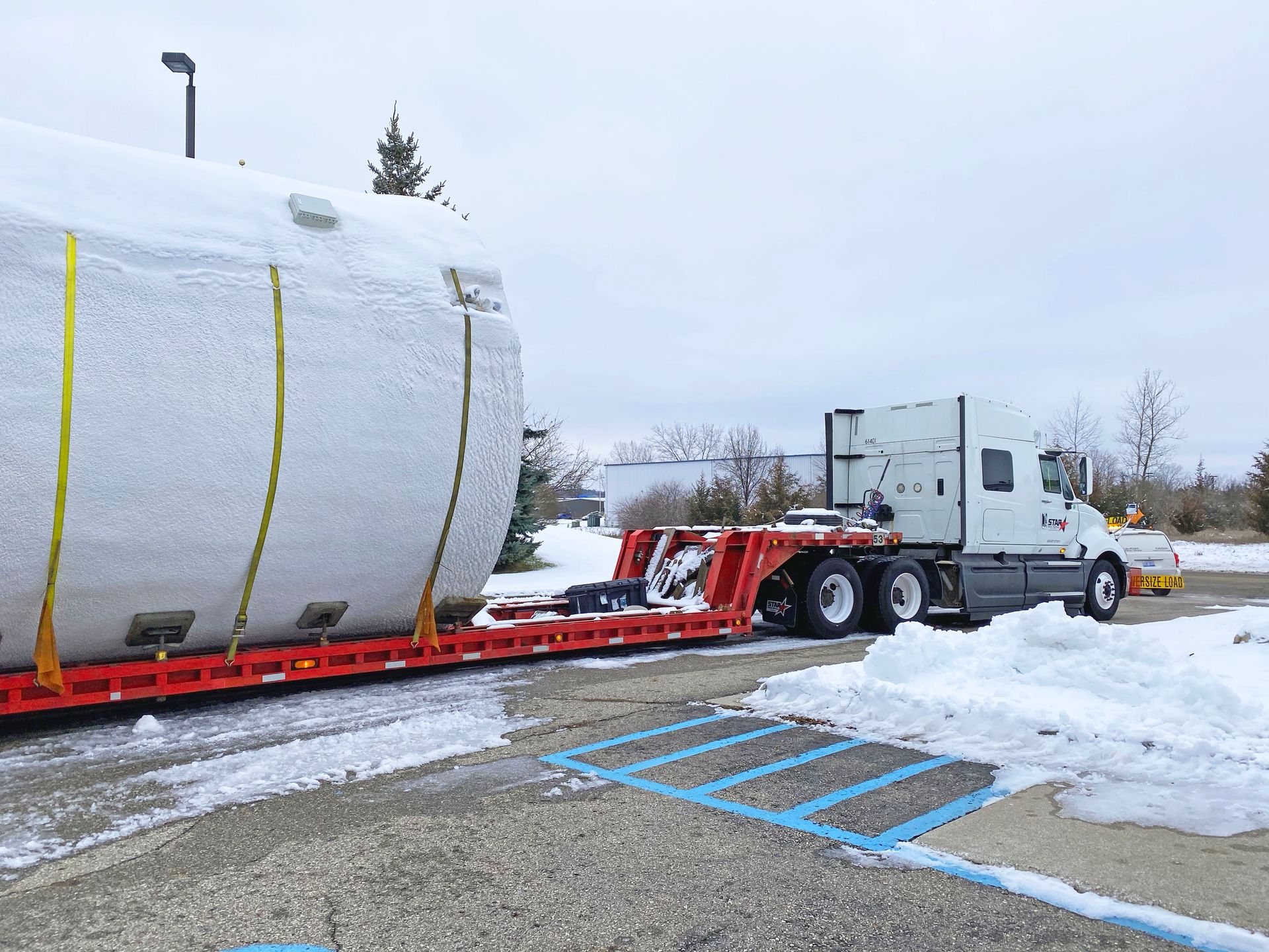 Truck hauling a large, cylindrical object covered in snow; parked in a snow-covered parking lot.
