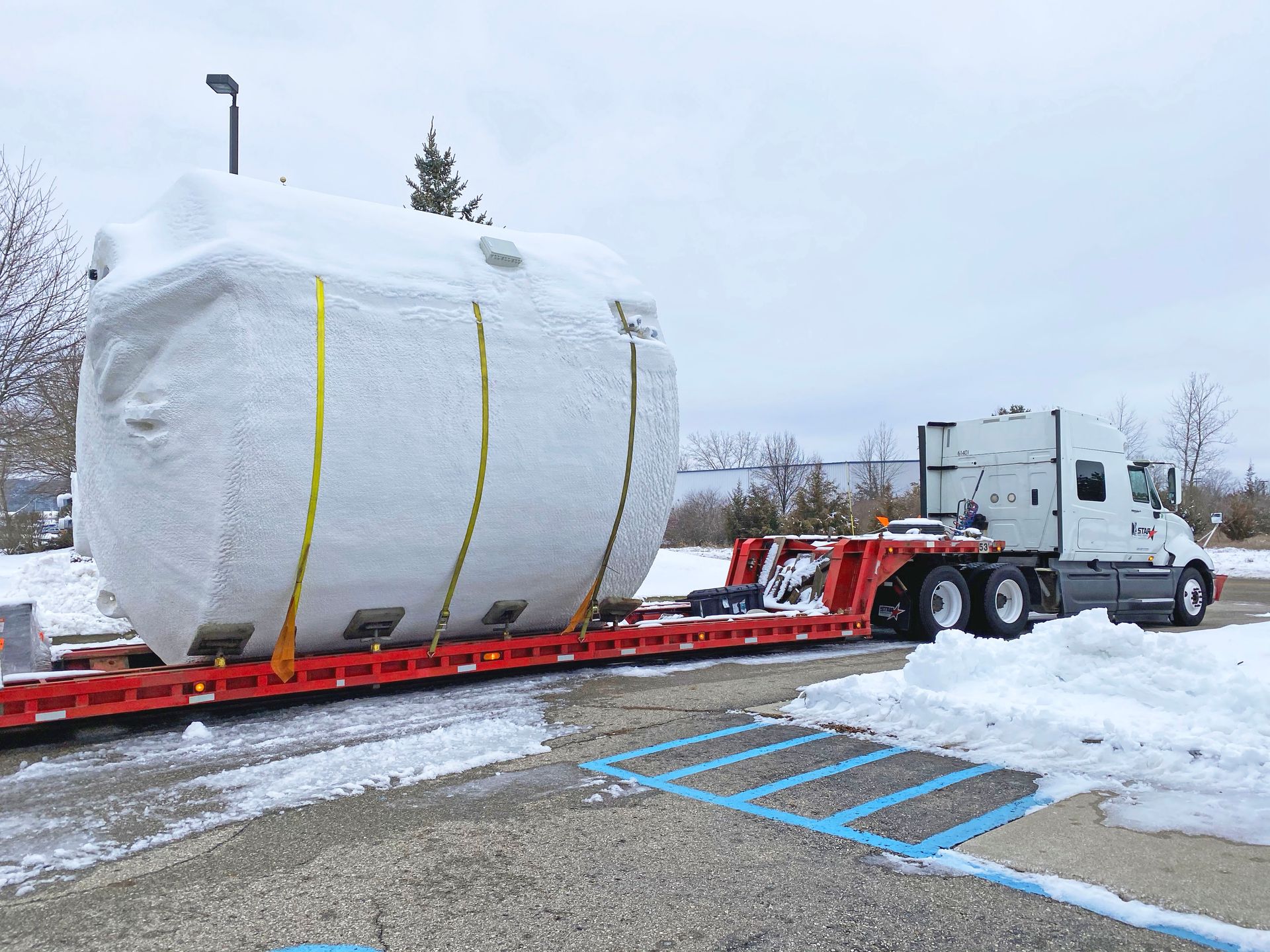 A large, wrapped object secured on a flatbed trailer, pulled by a semi-truck in a snowy parking lot.