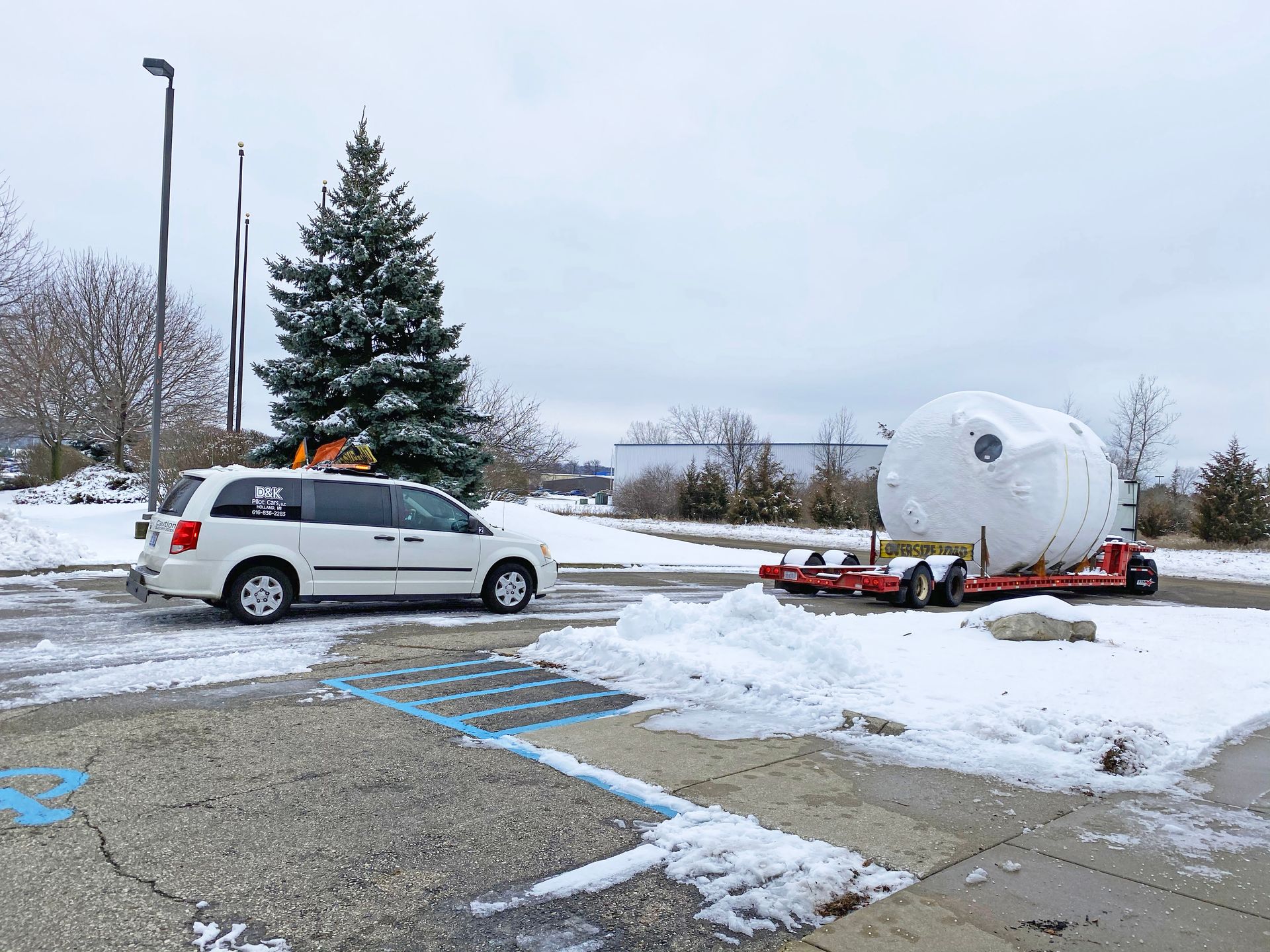 White van towing a large, wrapped, round object on a trailer in a snowy parking lot.