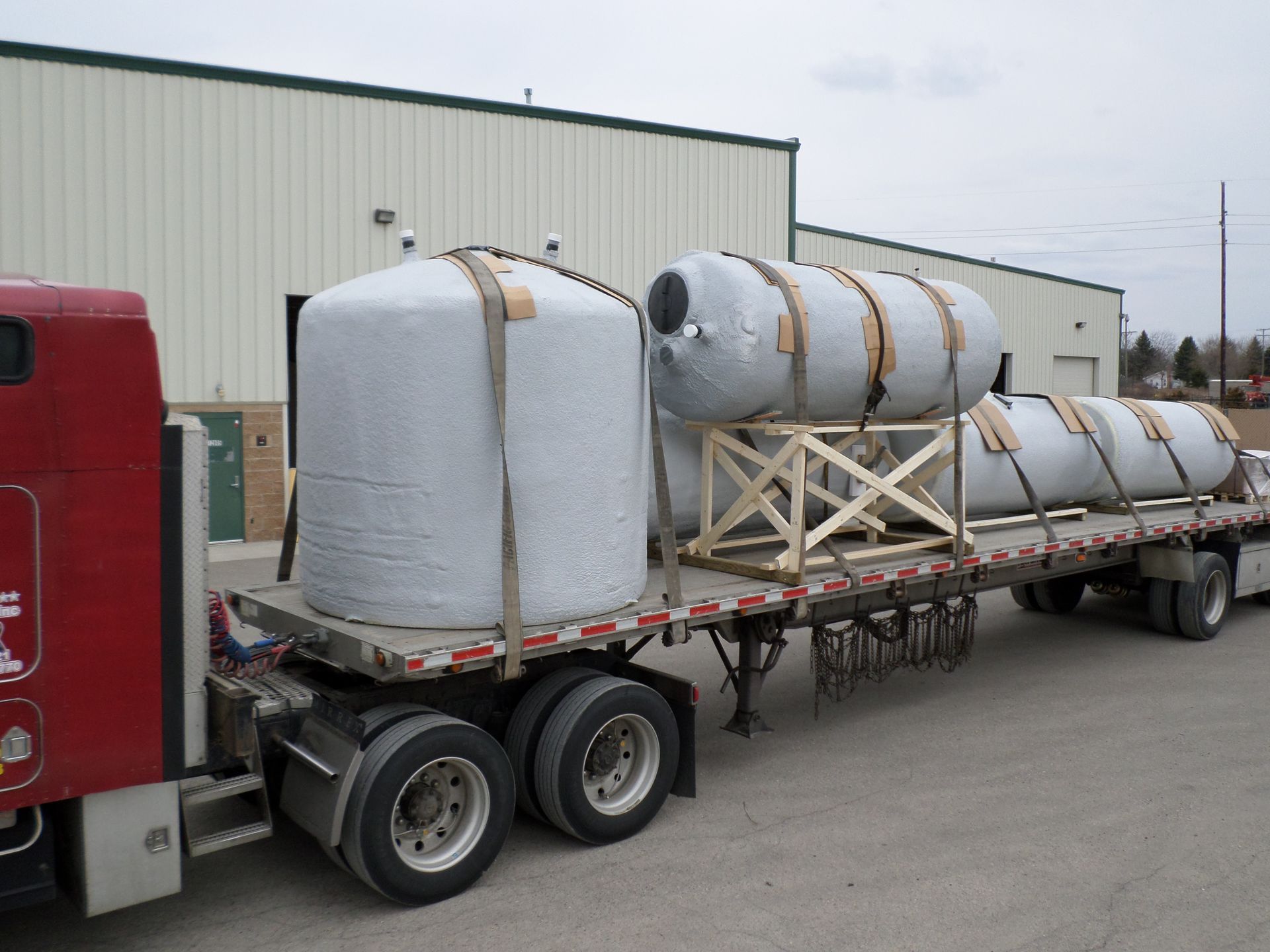 Truck carrying large, gray tanks secured with straps on a flatbed trailer, parked outside a building.