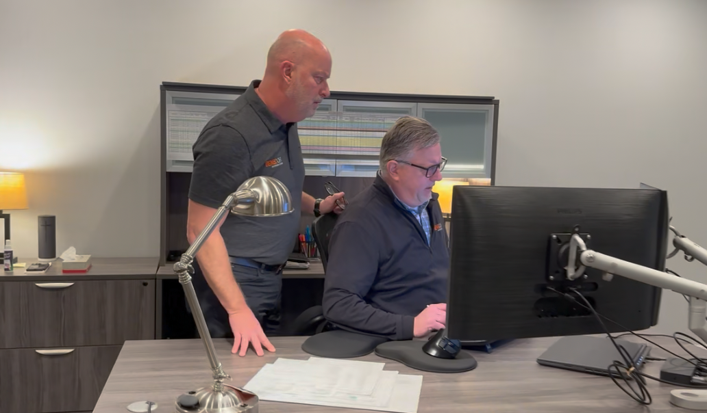 Two men at a desk; one standing, looking at the other who is at a computer. Office setting.