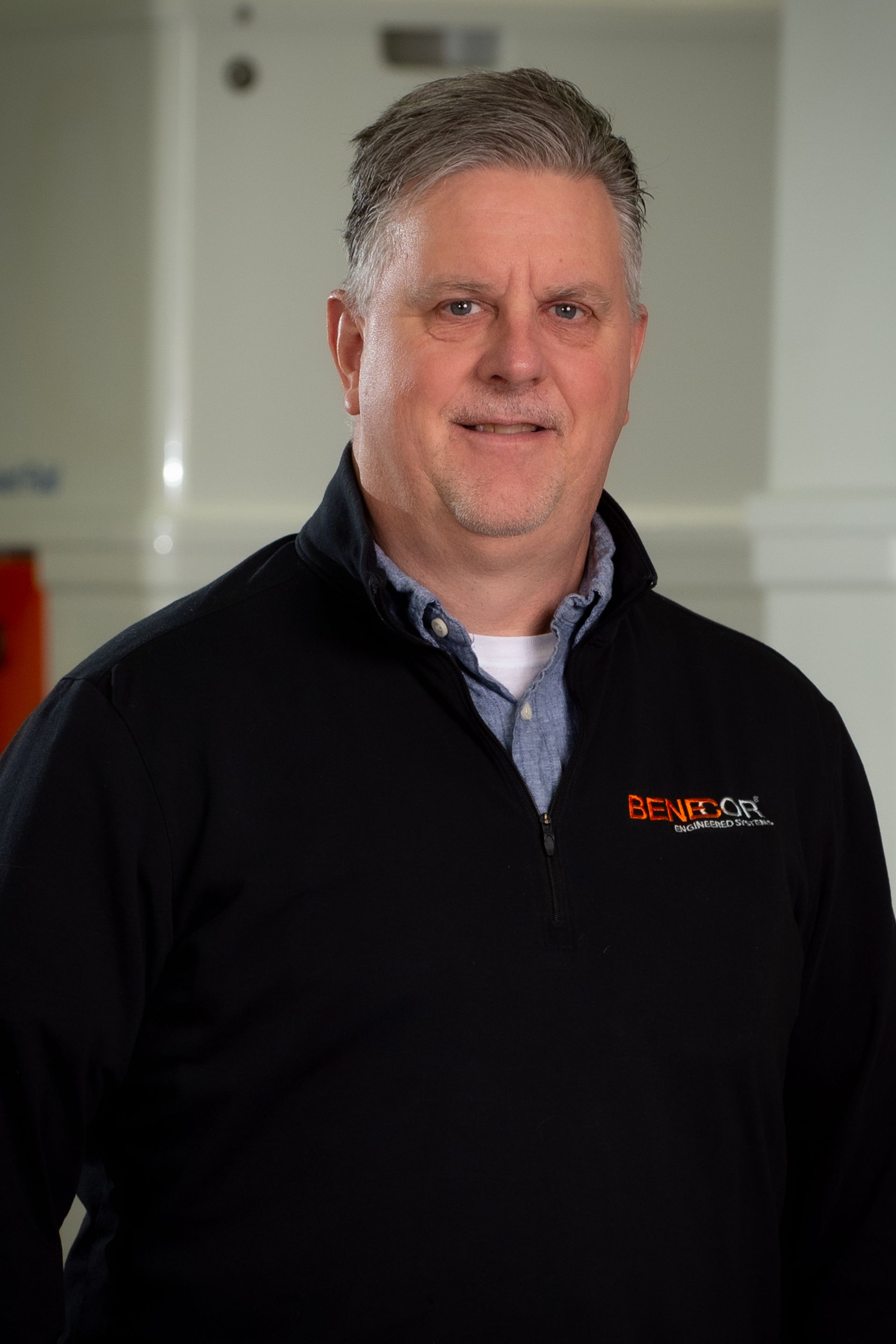 Man in a black jacket with logo, standing and smiling. Indoors.