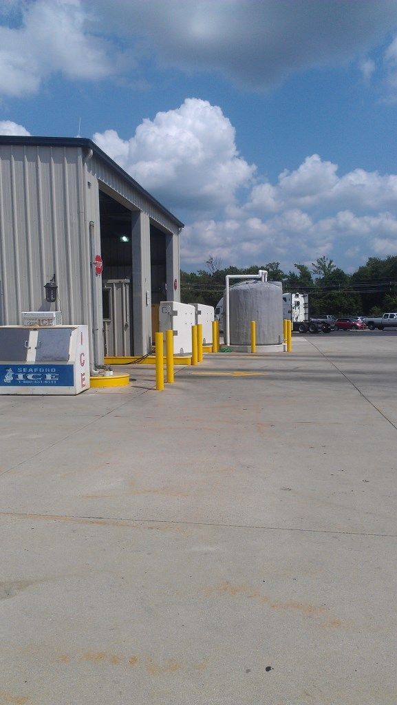 Exterior of a commercial building with equipment and storage tanks on a concrete pad under a partly cloudy sky.