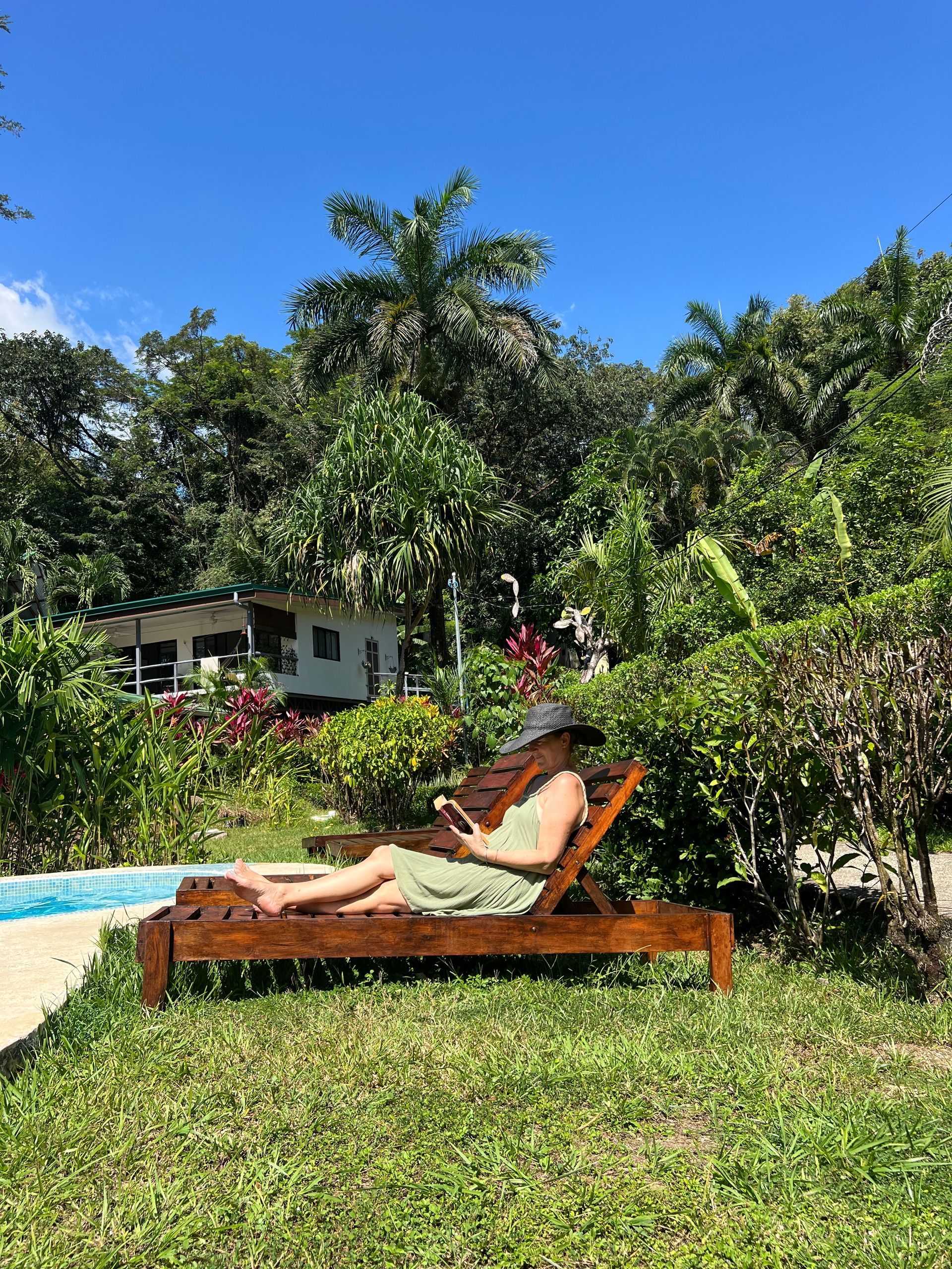 A woman is sitting on a lounge chair in the grass near a pool.