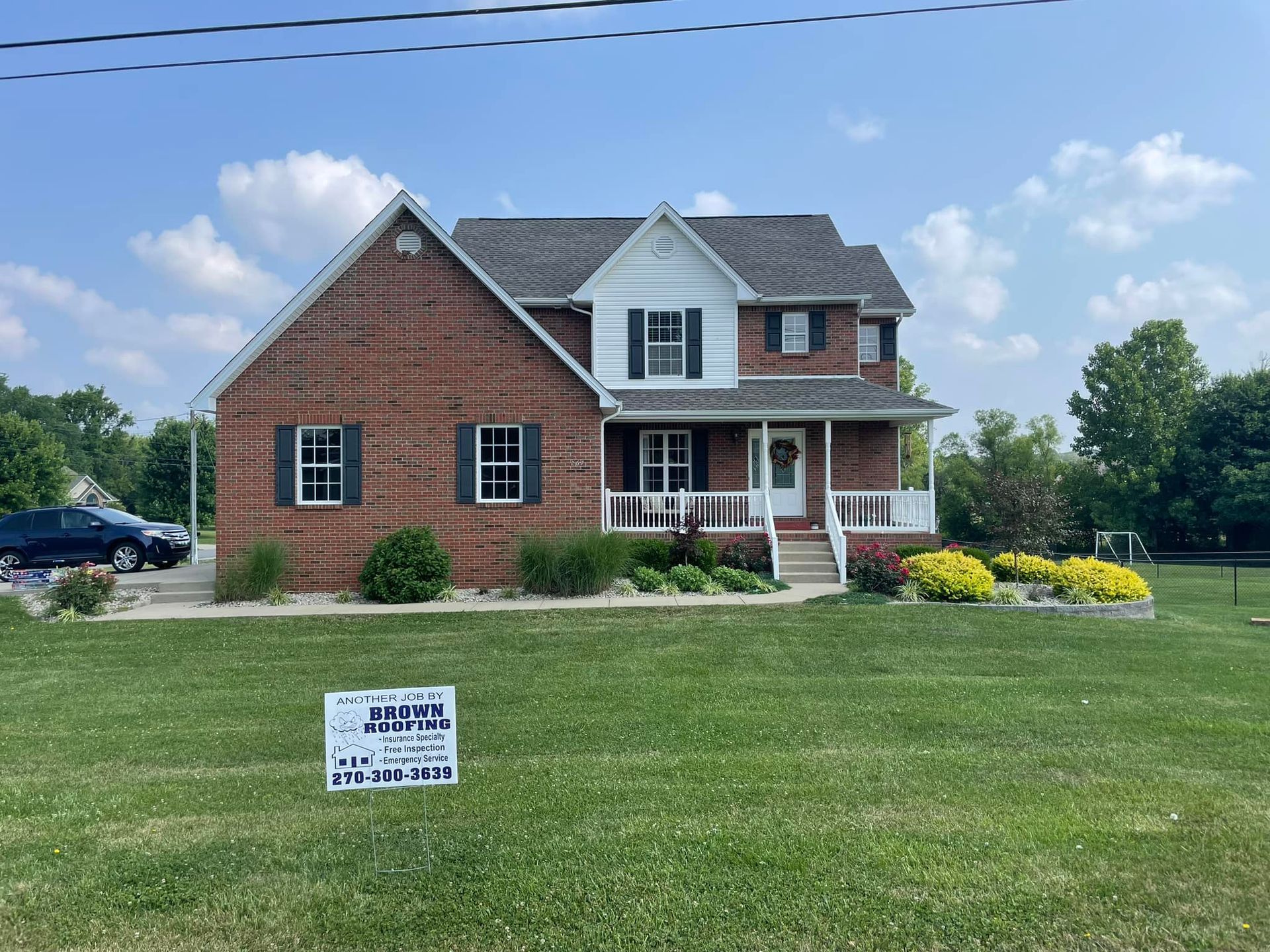 Brick house with white trim, dark shutters, and a front porch, on a grassy lawn under a partly cloudy sky.