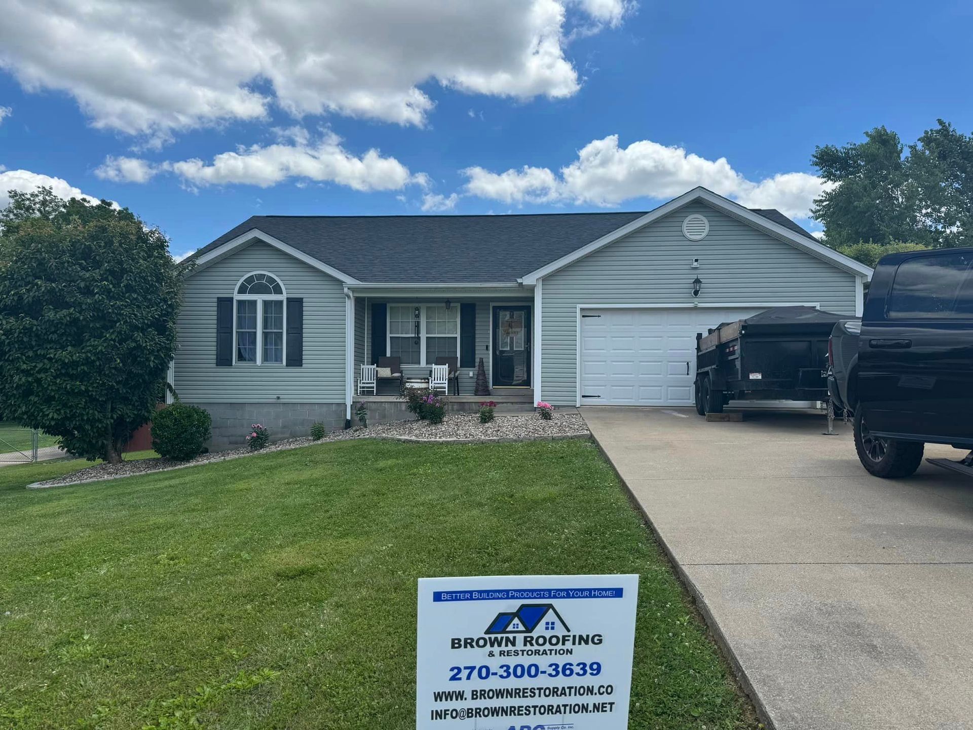 House with blue siding and new black roof; a sign indicates roofing company on lawn.