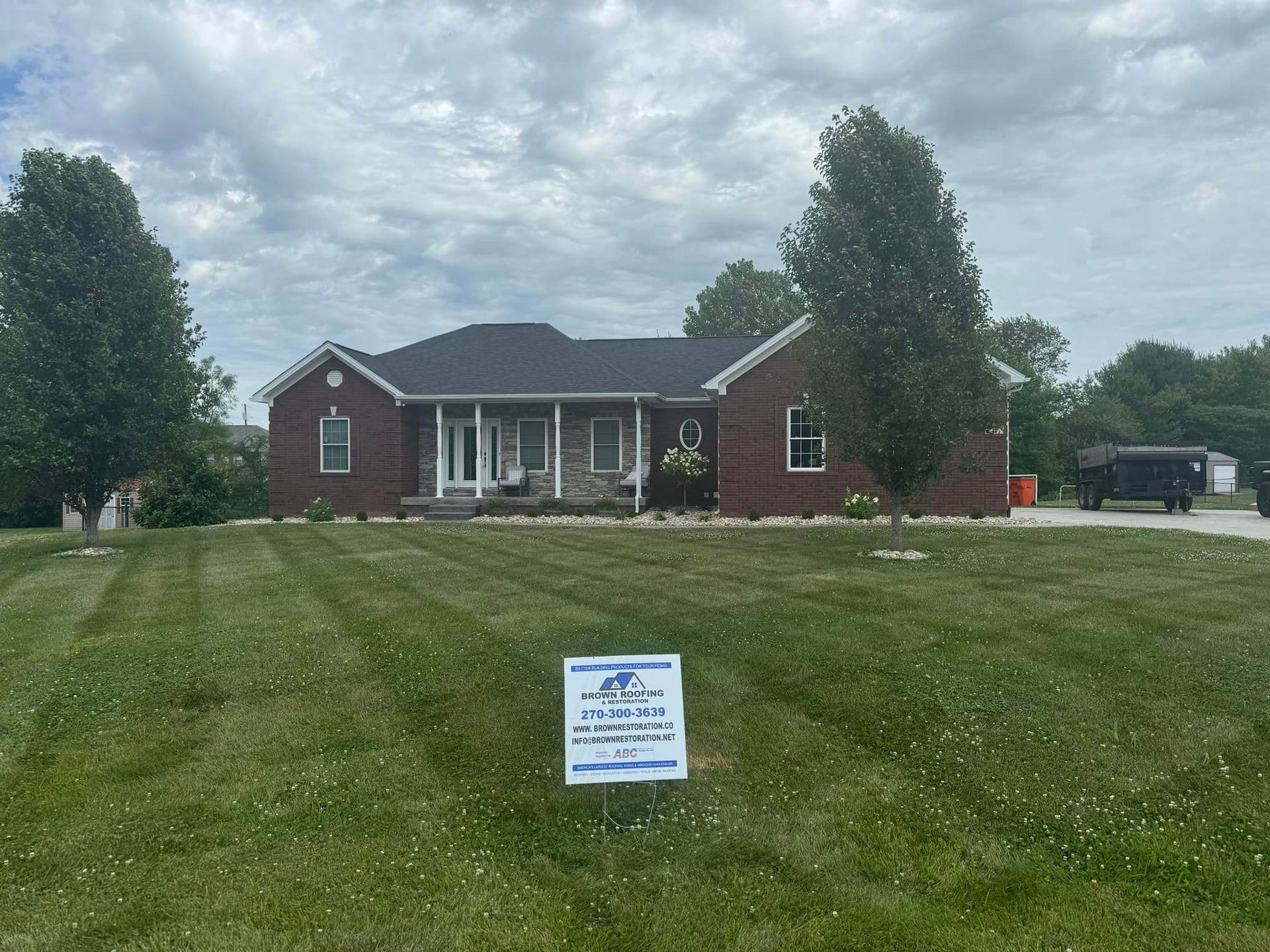 Brick house with black roof, trimmed lawn, and sign in yard. Trees flank the home. Overcast sky.