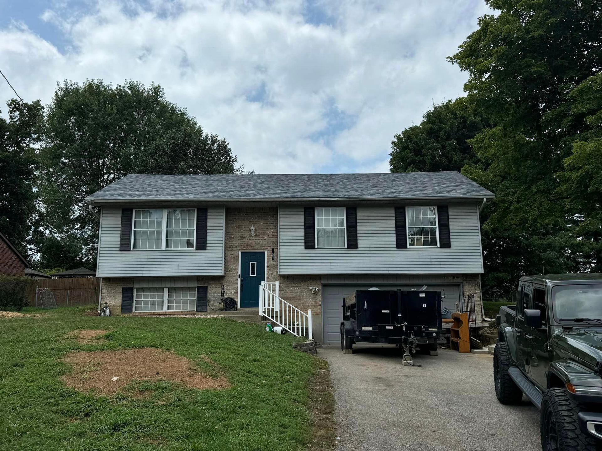 Split-level house with grey siding, blue door, and black shutters, parked trailer and jeep in driveway.