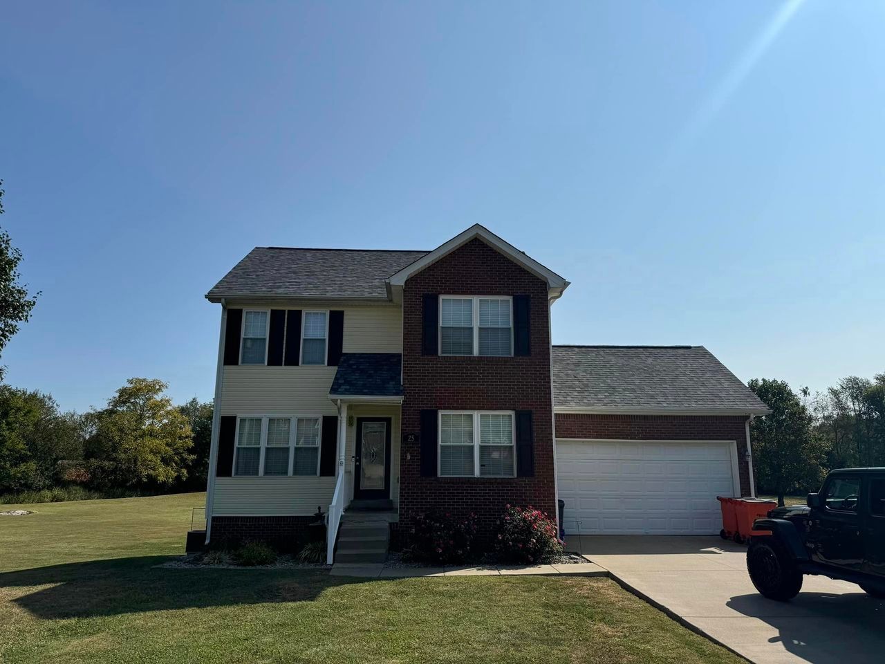 Two-story house with tan siding, brick facade, black shutters, and attached garage under a blue sky.