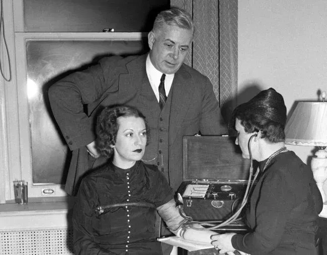 Man watches as a woman's arm is examined with a device by another person wearing a hat. Indoors.