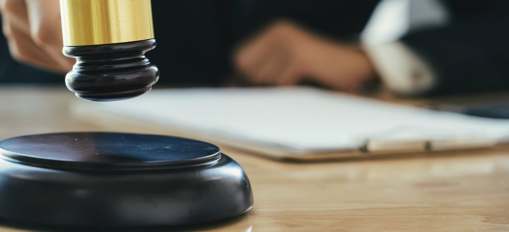 A gavel striking a wooden block on a desk, with documents and a person in the background.