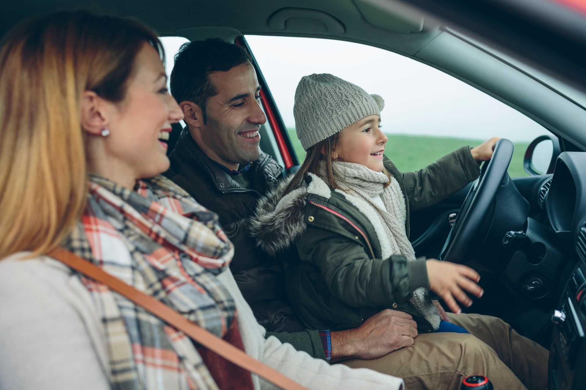 Family smiling in a car, child at the steering wheel, outdoors.