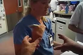 Nurse in blue scrubs talking with hands raised in a hospital room.