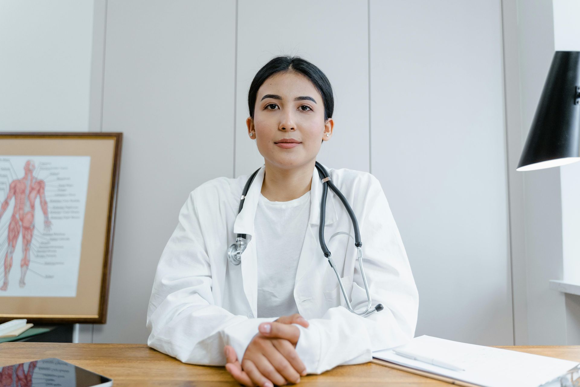 Doctor in white coat, stethoscope, sitting at desk, anatomical chart in background, indoor.