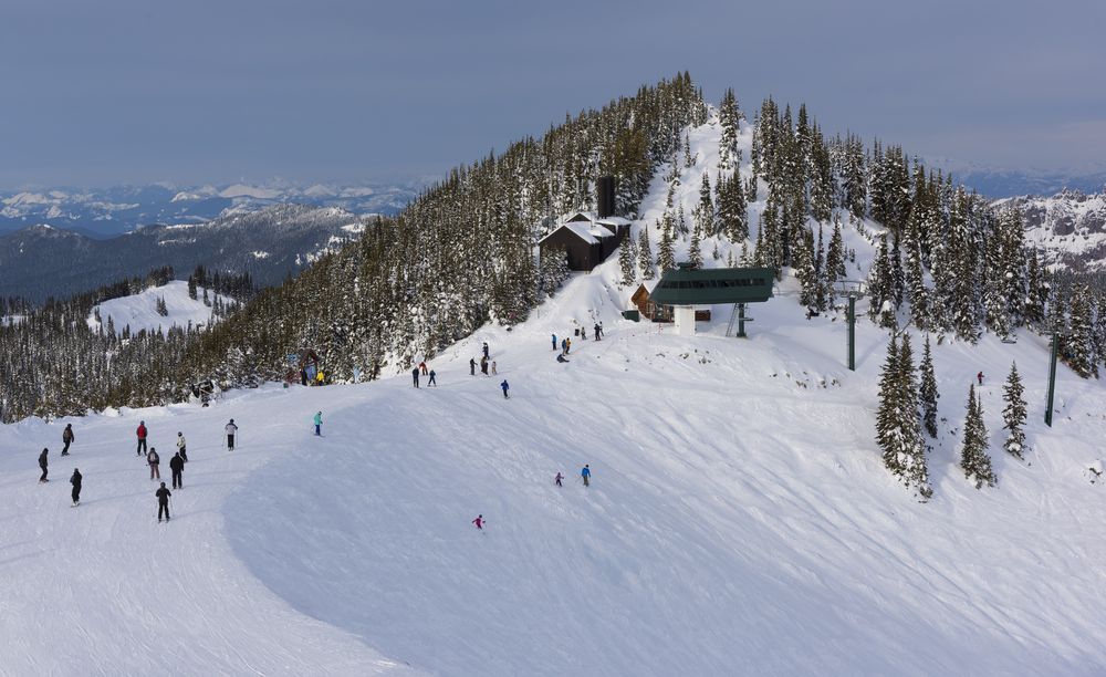 Snowy mountain slope with skiers and snowboarders, ski lift, buildings, and trees. Overcast sky.